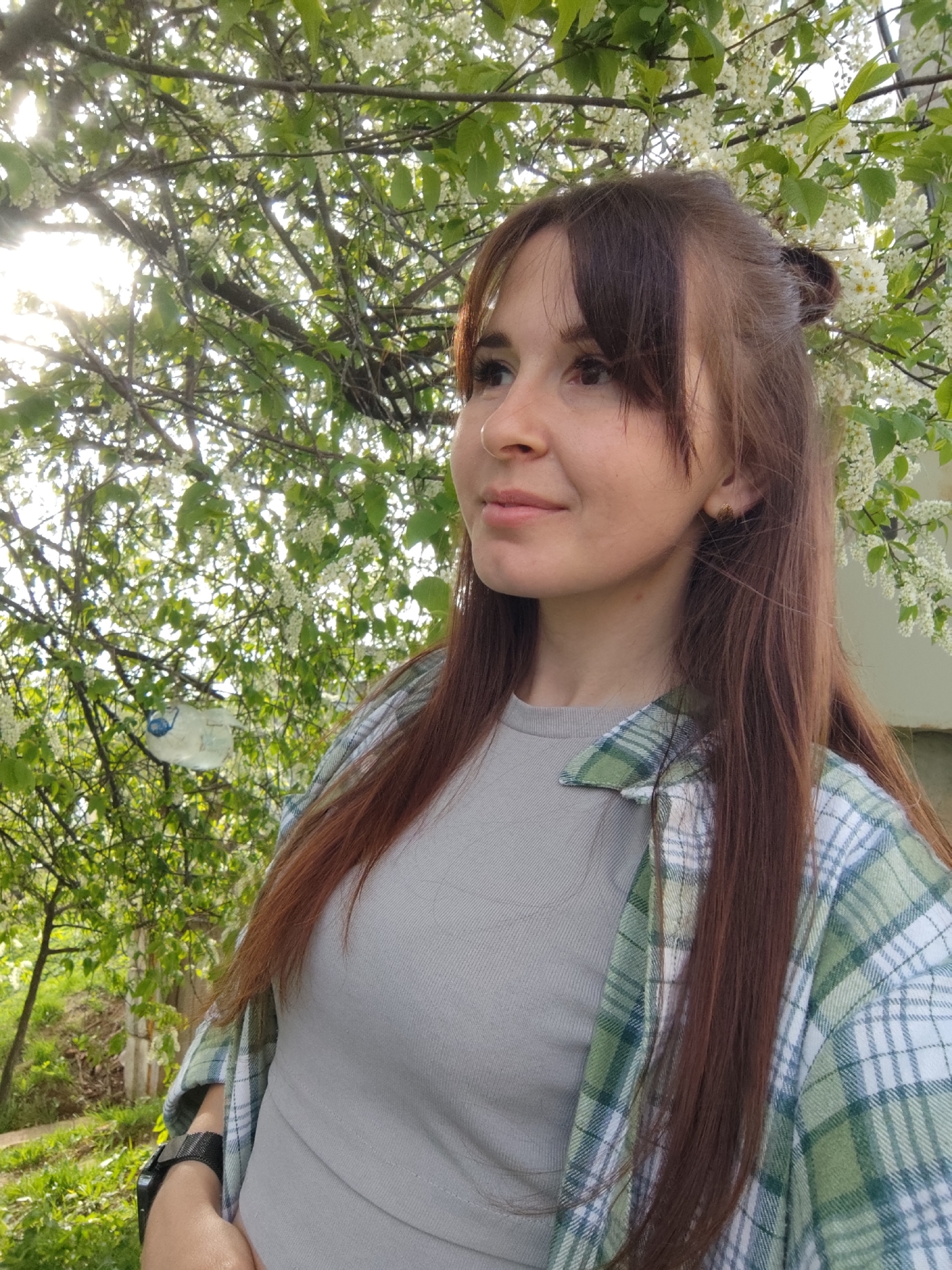 A woman stands outdoors, wearing a light-grey tee-shirt and a green and white plaid shirt. She has brown hair, with bangs and some of her hair pulled up into a bun. The background is filled with greenery, branches, and white blossoms. There is a blurred landscape with a grassy area and a glimpse of a structure in the background. The sun shines through the branches, creating a bright spot in the upper left. There are no indications to establish that this is in Фоки, Russia.