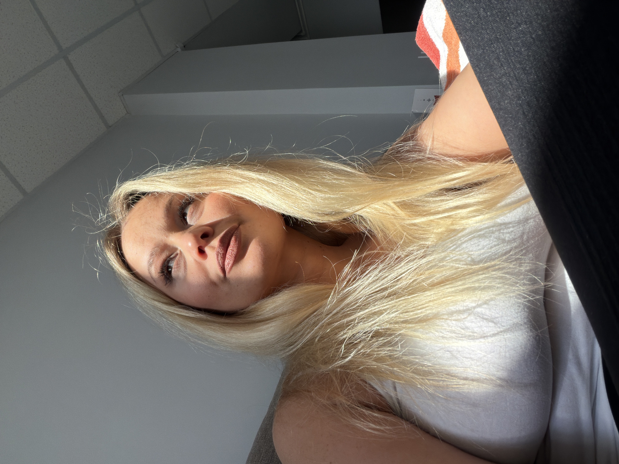 A woman with long blonde hair is pictured from a low angle, appearing to look down at the camera. She is wearing a white top. The background includes a ceiling with square panels and a grey wall. The location is not identifiable as Sarapul, Russia.