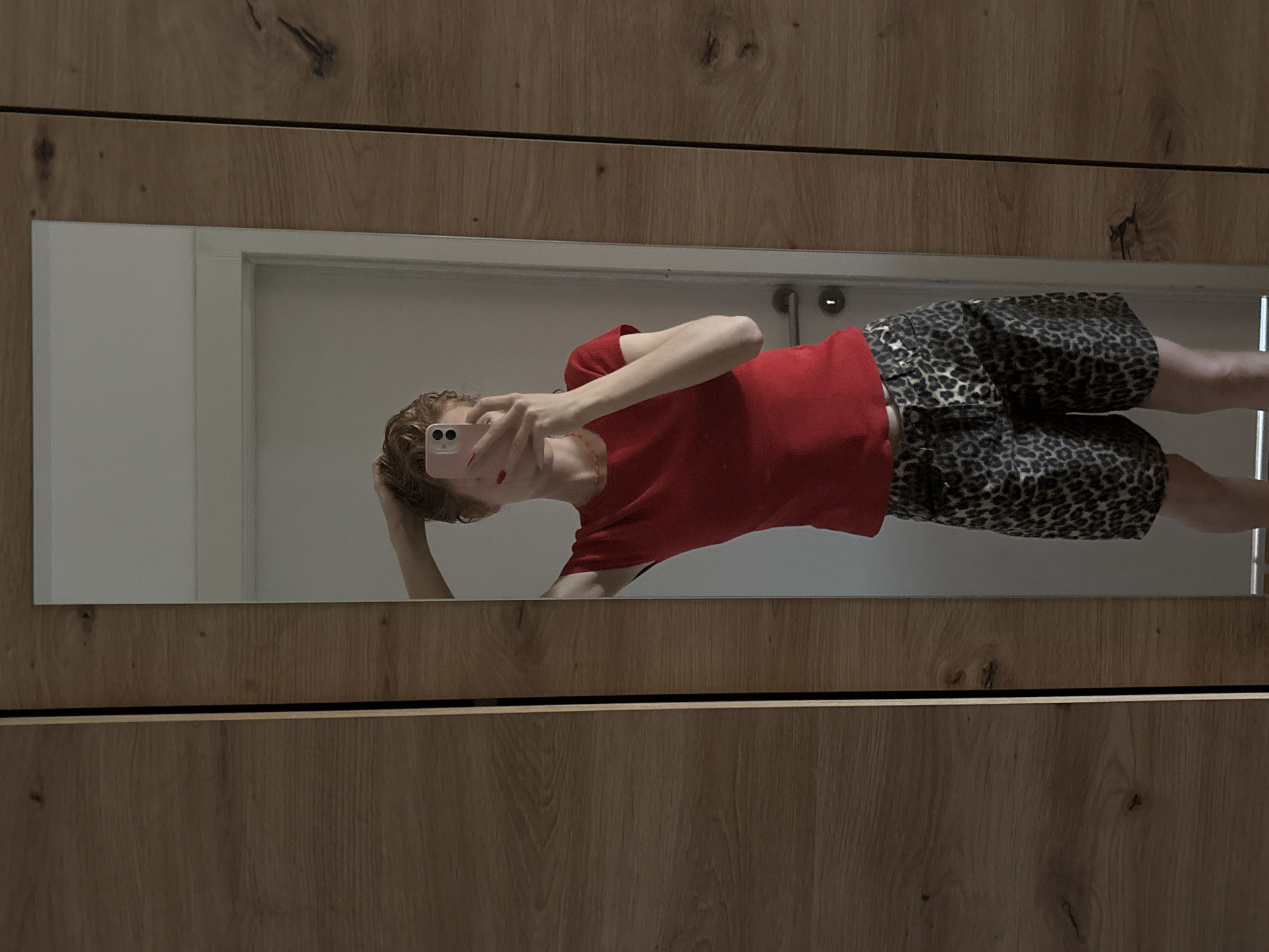 A person with short, reddish-blonde hair is standing in front of a full-length mirror, taking a selfie with a pink smartphone. They are wearing a red t-shirt and leopard-print shorts. The person's left arm is raised, with their hand in their hair. They are reflected in the mirror, which is positioned next to a wooden wardrobe. The background appears to be an interior space with a white door visible in the reflection. There is no indication of the city of Kragujevac, Serbia in the image.