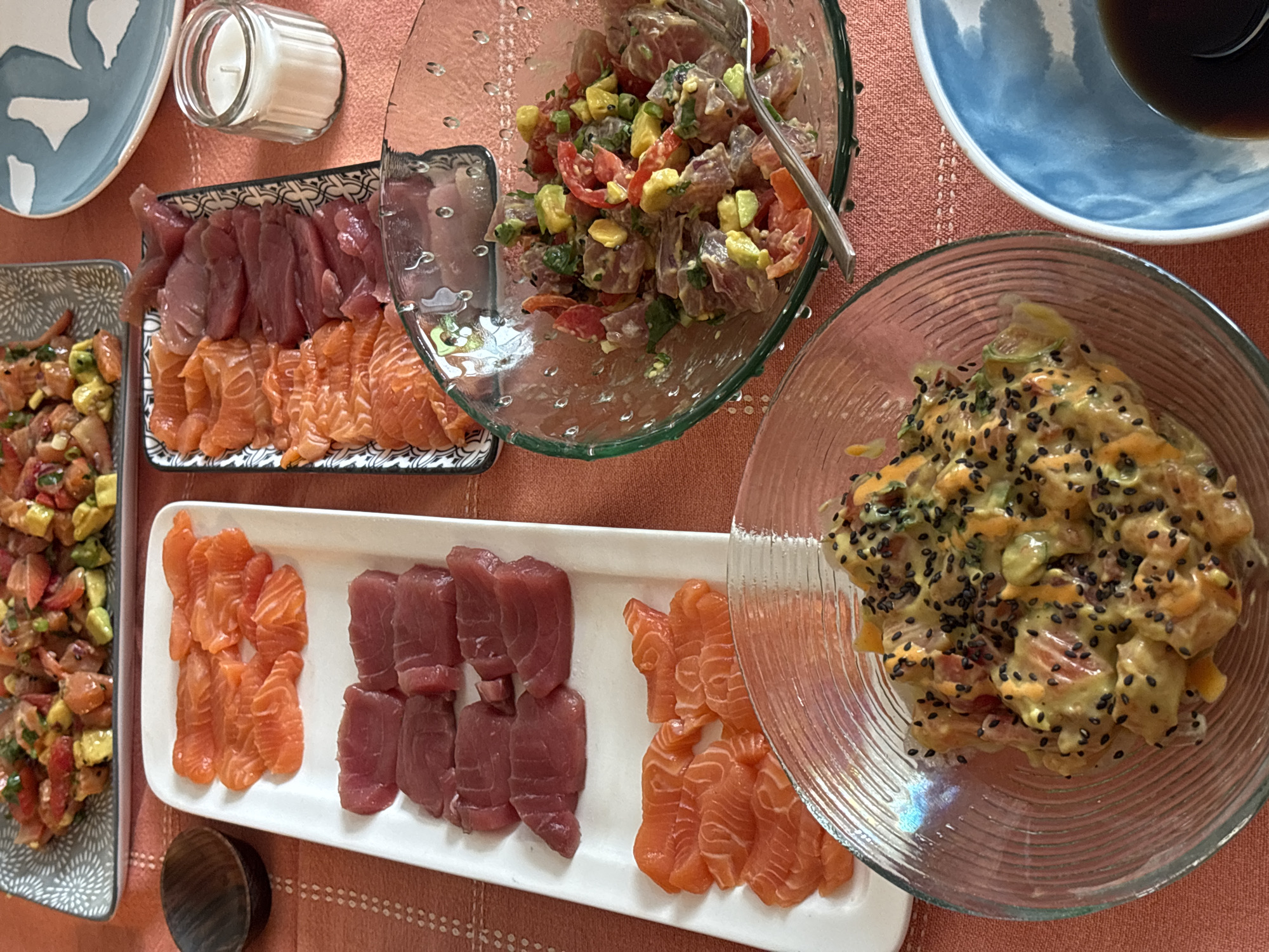 A table is set with several dishes of food, likely in a residential setting. There are two long rectangular plates, each displaying raw fish, possibly tuna and salmon. One is arranged with salmon on the left and tuna on the right; the other has the arrangement reversed. There are also two bowls of mixed ingredients, possibly poke, with vegetables and fish. A small candle sits to the left of one rectangular plate. A bowl of soy sauce is also visible. The location is not discernible from the image.