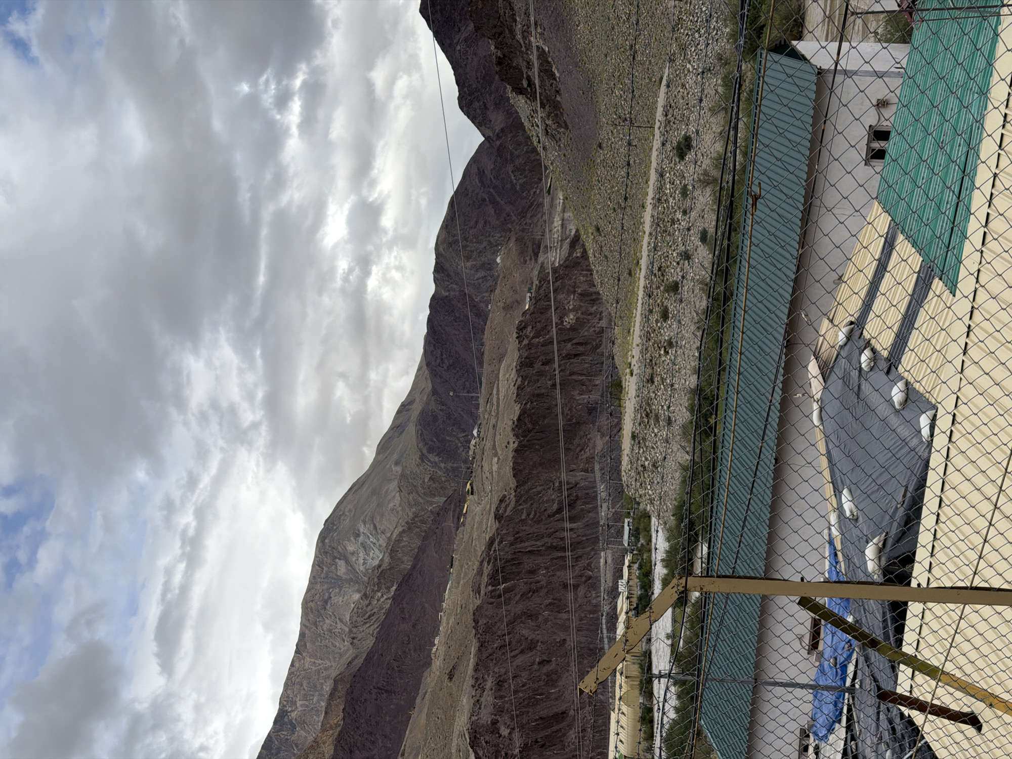 The image shows a mountainous landscape with a cloudy sky. In the foreground, there is a fence and some buildings with green and yellow roofs. Beyond the buildings, a river can be seen, and the mountainous terrain rises steeply. Some of the mountains appear to be bare rock, while others are covered in sparse vegetation. There are also some structures visible on the mountainside, possibly houses or military outposts. The scene suggests a remote, possibly rugged location. The image is taken in Kiari, India.