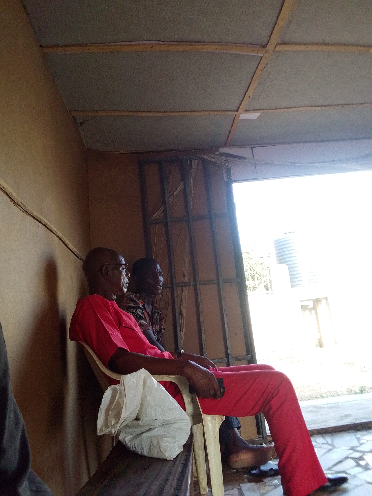 Two men are sitting in a room. The older man, wearing a red shirt and pants, is seated in a white plastic chair, with a white plastic bag on the bench to the side. The other man is in a patterned shirt. There is a metal grate-like structure near the doorway. Outside, a water tank is visible. The location is possibly in Lokoja, Nigeria based on the prompt. The room has a clay wall and a tiled ceiling.