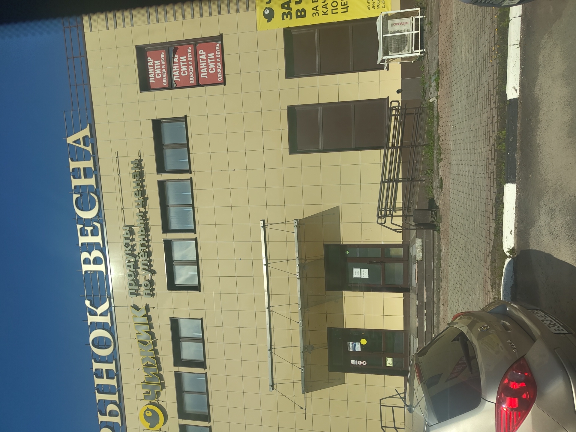 The image shows the facade of a commercial building with a large sign at the top that reads "РЫНОК ВЕСНА" (Spring Market). Below this, a smaller sign features a yellow bird logo and the text "ЧИЖИК ПРОДУКТЫ ПО УЛЕТНЫМ ЦЕНАМ" (Chizhik Groceries at Insane Prices). To the right, there are three vertical red advertisements that read "ЛАНГАР СИТИ ОДЕЖДА И ОБУВЬ" (Langar City Clothing and Shoes). The building has a tiled facade, several windows, and two entrances with glass doors. A ramp for accessibility is visible in front of the entrances. An air conditioning unit is mounted outside the building on the right. In the foreground, the rear of a silver car is visible, parked on a paved area. The scene appears to be in a city, under a clear blue sky. Based on the presence of the "Chizhik" chain and the style of the building, it is likely located in the city of Ramenskoye, Russia.