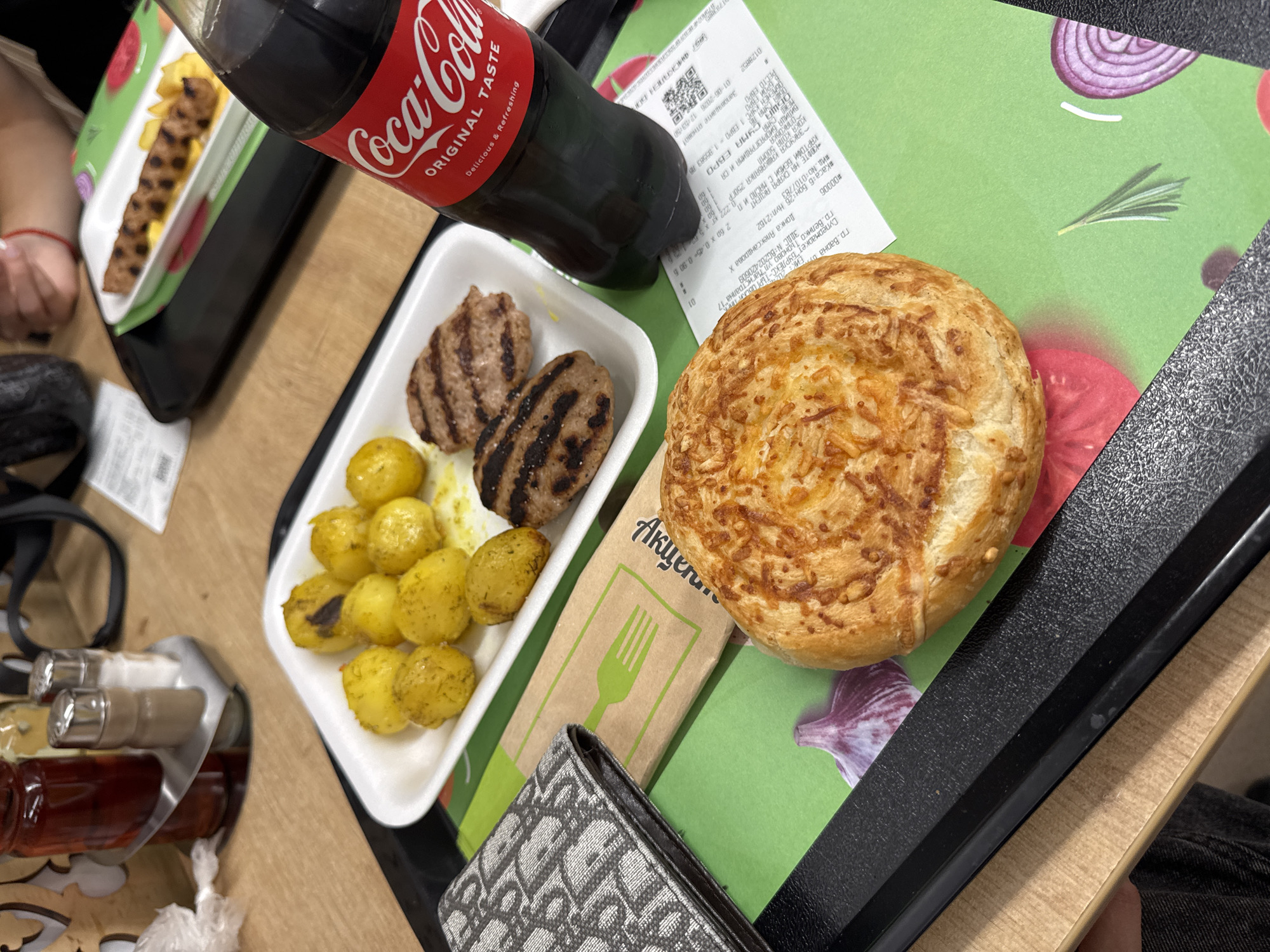 A high-angle shot shows a meal on a black tray atop a table. The location is in the city of Veliko Tarnovo, Bulgaria, specifically at the supermarket "Burlex-Targovski Tex" at 17 Magistralna Street.
The meal consists of two grilled patties, small boiled potatoes coated in oil, and a round cheese-topped bun. A bottle of Coca-Cola is positioned near the food. A receipt from the supermarket is partially visible, displaying items purchased including "Kartofi Baby s Maslo," "Kofte na Skara Akcent," "Zakuska Kashkavalka," "Coca-Cola 500ml," and "Vilica biorazgradima i Sa."
A Dior-patterned wallet rests near the food, and there are condiments in small bottles. The tray rests on a surface with a decorative pattern. Two hands and part of an outfit are visible.