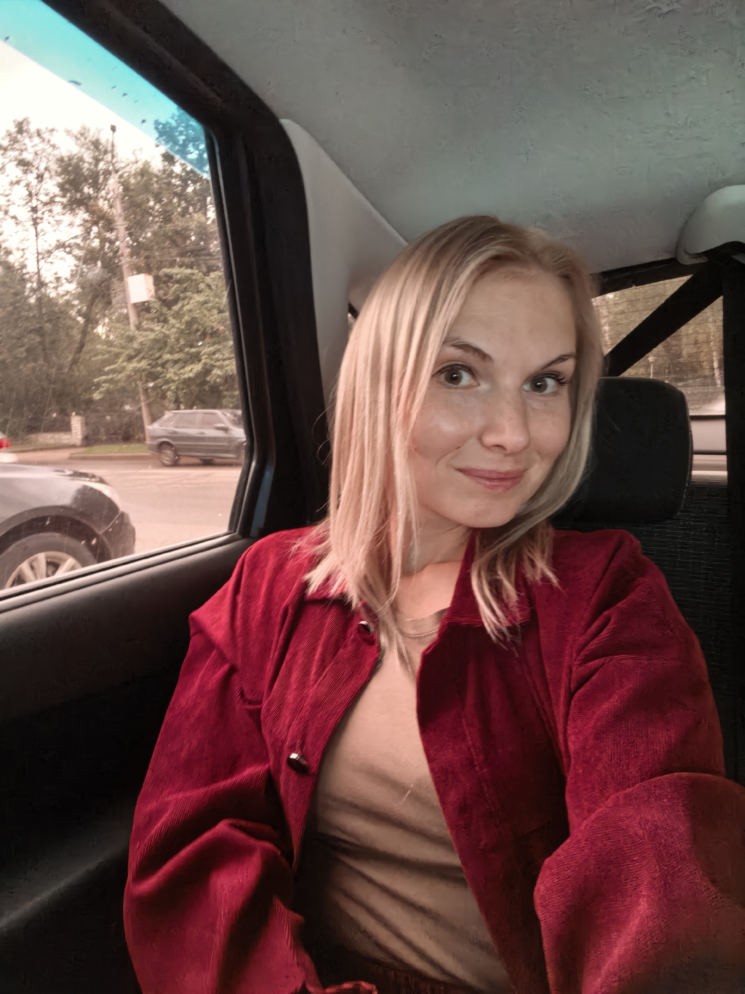 A young woman with blonde hair and fair skin is taking a selfie in the backseat of a vehicle. She is wearing a red corduroy jacket over a beige top. The interior of the car is visible, including the seat upholstery and the window. Outside the window, trees, a street, and another car can be seen. The scene appears to be in a residential area of Sterlitamak, Russia, given the presence of deciduous trees and a typical street scene. The lighting suggests it is daytime.