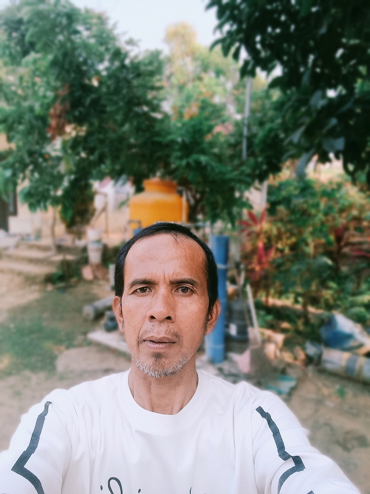 A middle-aged man with short, dark hair and a receding hairline is pictured in a selfie taken outdoors in Bulangan Barat, Indonesia. He has a light gray beard and mustache. He is wearing a white t-shirt with dark stripes on the sleeves. The background is blurred but appears to be a yard with trees, some with green foliage and others with red and green leaves. A large yellow water tank is visible behind the man's head to the left. The ground is dirt and gravel.