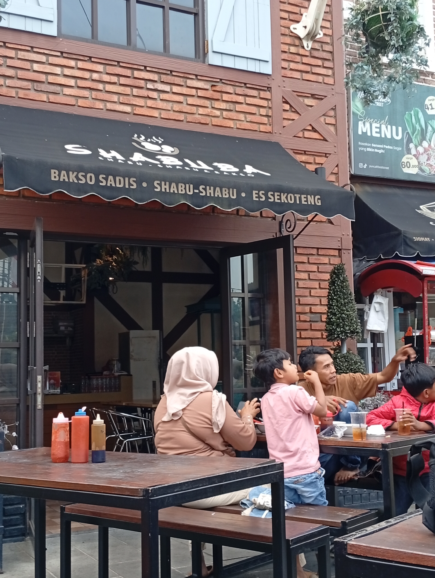 The image depicts an outdoor seating area of a restaurant in Cimacan, Indonesia. A black awning above the entrance displays text that includes "BAKSO SADIS", "SHABU-SHABU", and "ES SEKOTENG". Several people are seated at wooden tables. In the foreground, a wooden table holds three condiment bottles: two red and one brown with a blue lid. To the right, a man in a brown t-shirt is pointing, seemingly interacting with a young boy in a pink shirt and blue jeans who is also seated at the table. Another child in a red jacket is visible to the right, holding a drink. A woman wearing a light brown hijab and a matching top is seated at another table to the left. The restaurant building features brickwork and timber framing. A sign with "Special MENU" is visible on the right side of the building. The overall scene suggests a casual dining environment.