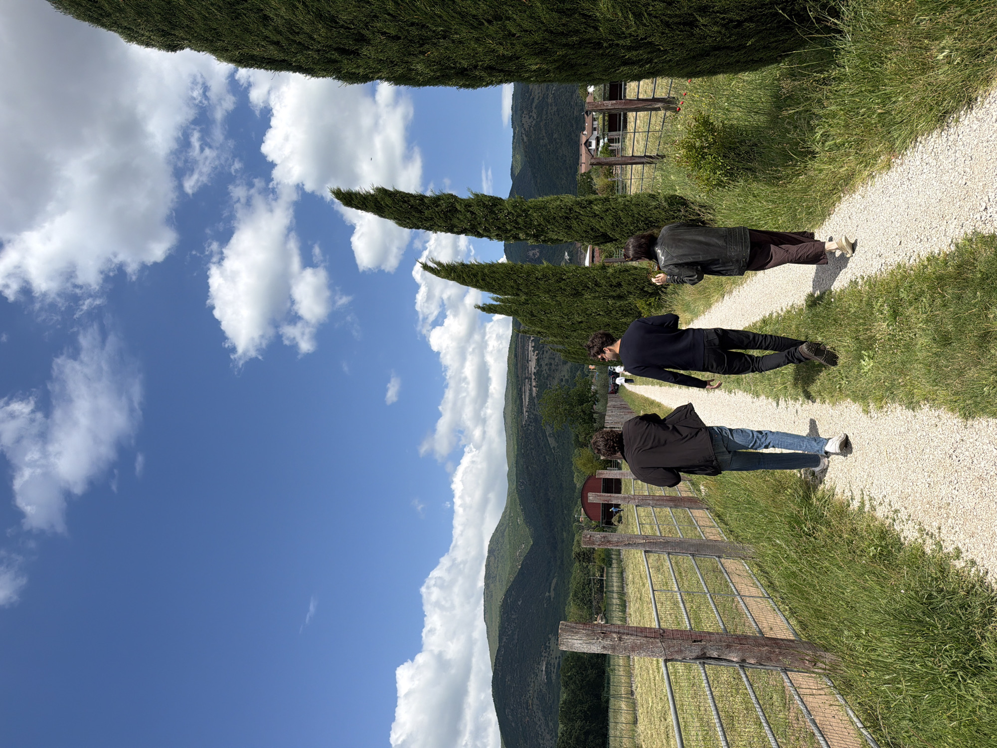 Three people are walking on a gravel path in Sala Consilina, Italy. The path is bordered by green grass and a wooden fence on one side, and tall, thin trees on the other. Behind the trees is a mountain range. The sky is blue with white, puffy clouds. The first person is wearing a black jacket, brown pants, and light-colored shoes. The second person is wearing a black jacket and black pants. The third person is wearing a black jacket, blue jeans, and gray shoes.