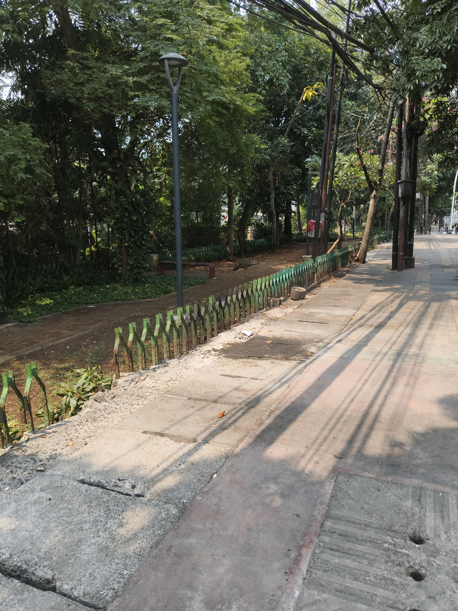 This image depicts a sun-dappled pedestrian walkway adjacent to a park area in South Jakarta, Indonesia. A row of green metal fence posts runs along the edge of the park, separating it from the sidewalk. The park itself is lush with trees and vegetation. A lamppost stands near the fence, and power lines are visible overhead, strung between poles. The sidewalk is made of concrete slabs, some of which appear worn or cracked, and is casting long shadows from the surrounding trees. The overall scene suggests a public urban green space.