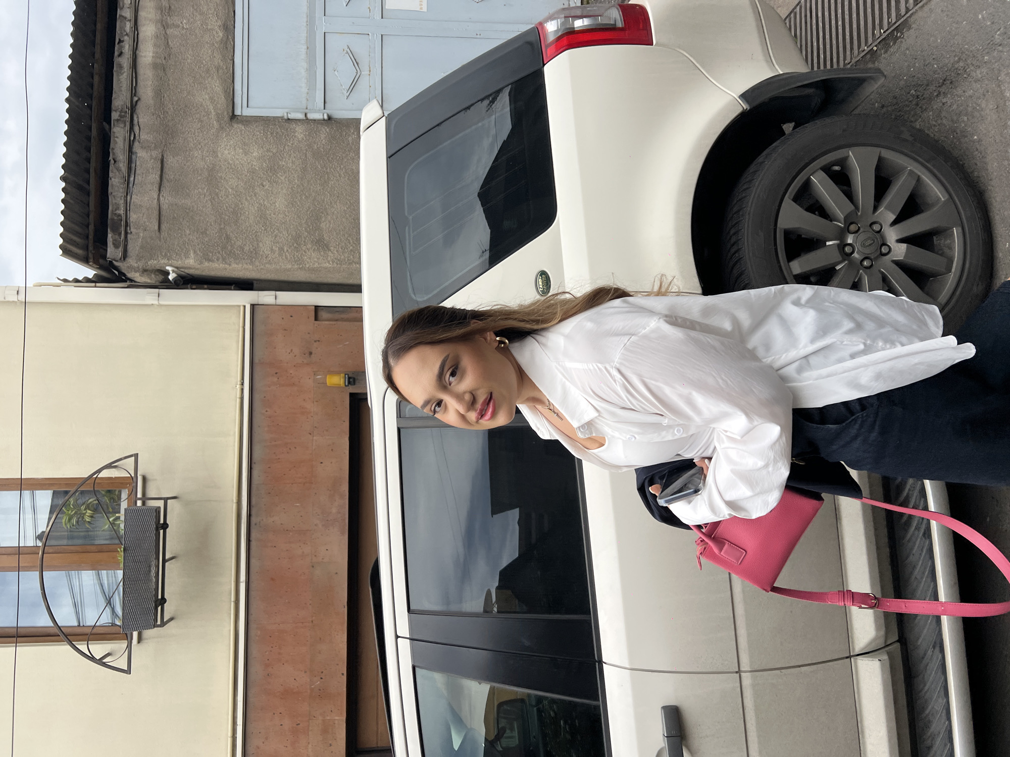 A medium, eye-level, full shot captures a woman with long brown hair, fair skin, and light makeup, wearing a white button-down shirt and dark jeans. She's holding a phone and a bright pink purse. She stands next to a white Land Rover, with a grey alloy wheel partially visible.
The location appears to be an urban street in Yerevan, Armenia. The backdrop features weathered buildings with brown and tan facades. A grey building with a corrugated roof and a blue metal door is partially visible at the top of the frame. A decorative metal window grate is seen on the left side of the image. The lighting is overcast, suggesting an urban environment on a cloudy day.