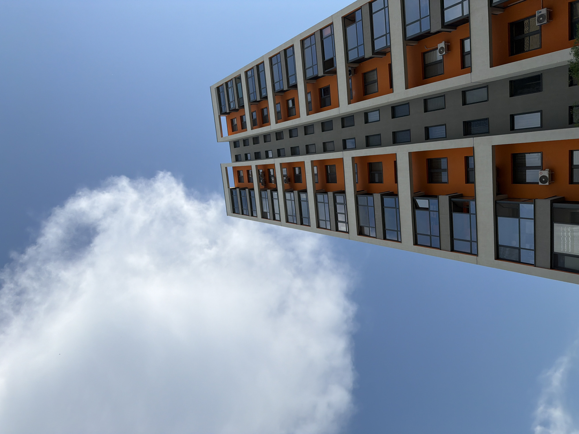 The image shows a tall building with a modern architectural style, featuring alternating sections of orange and grey panels, and numerous windows. The building's orientation gives a worm's-eye view, with a blue sky and a large white cloud visible. The architectural style and colors suggest a contemporary residential or commercial structure. Based on the metadata, this location is in Tyumen, Russia.