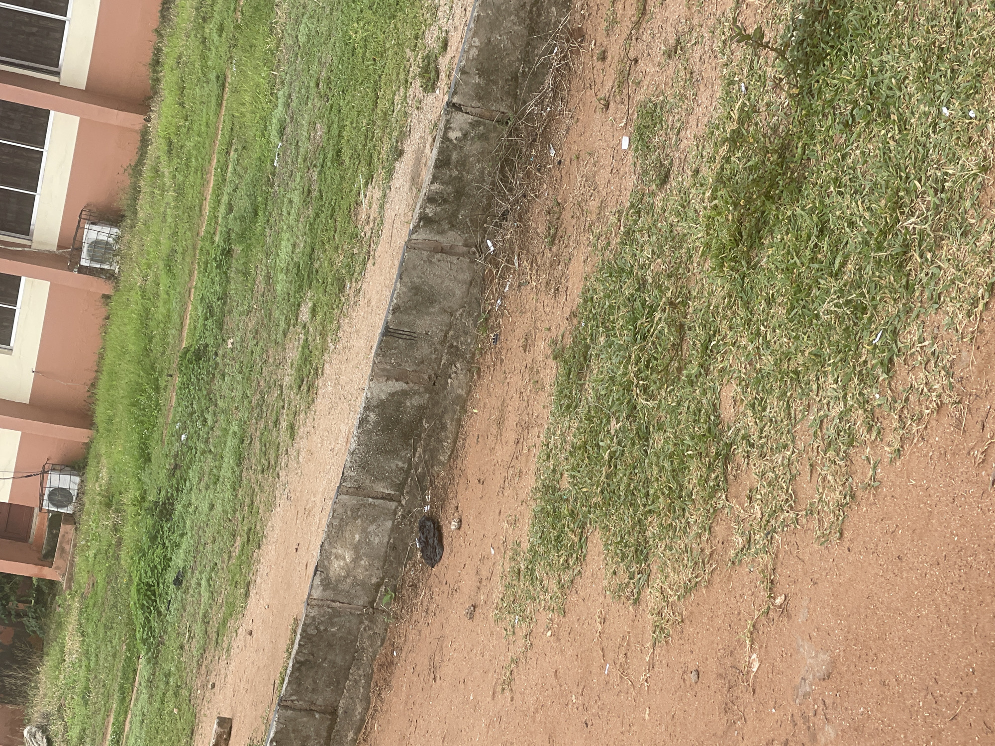 The image displays an outdoor scene in Lokoja, Nigeria, likely in a public area or near a building. The foreground consists of reddish-brown soil with patches of dry, light green grass. A low concrete barrier runs horizontally across the midground, separating the soil area from a strip of overgrown grass and vegetation. Beyond this, a building with light-colored walls and a reddish-brown trim is visible, with windows along the upper section. An air conditioning unit, enclosed in a metal cage, is mounted on the wall near the ground. There are some scattered pieces of debris, including a dark, crumpled object that appears to be a plastic bag, and a few small, light-colored fragments. No people are present in the image.