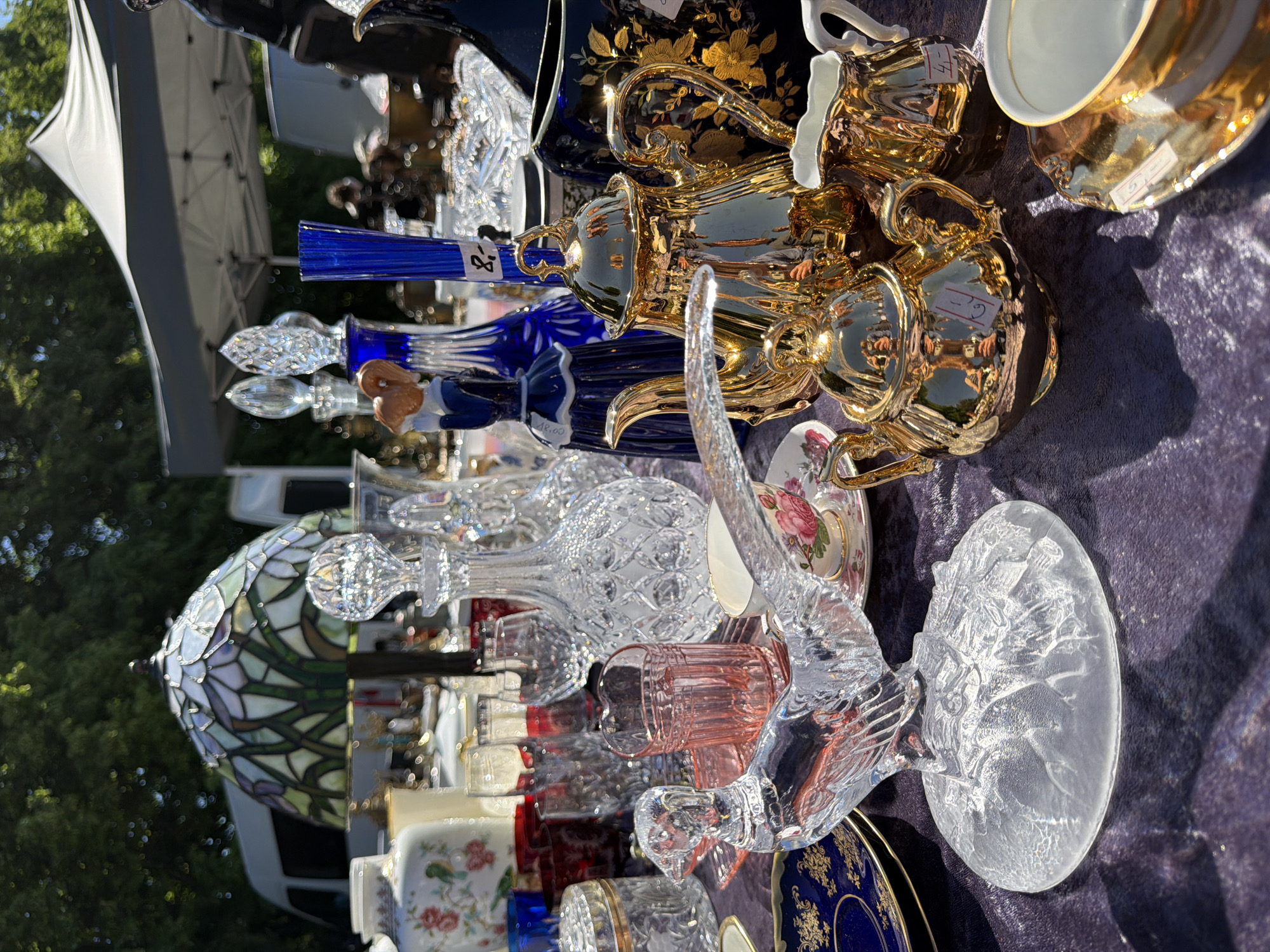 The image captures a close-up, eye-level perspective of a vendor's stall at an outdoor market, featuring an assortment of decorative objects. The scene is illuminated by direct sunlight, casting distinct shadows on the display surface. The primary subject is a table covered with a textured, dark purple fabric. Arranged on this surface are numerous items, predominantly crystal glassware, gold-toned metallic objects, and porcelain. **Specific objects visible include:** * **Glassware:** * A prominent clear crystal decanter with a diamond-cut pattern and an ornate stopper, positioned centrally. * A clear glass figurine of a stylized bird, possibly a pheasant or grouse, resting on a frosted oval base, located in the lower-left foreground. * A tall, ribbed blue glass decanter or vase with a stopper, situated in the mid-left. * Several clear drinking glasses and tumblers, some with etched or cut patterns, and a single pink ribbed glass tumbler are visible behind the glass bird and beside the decanter. * A clear glass jar with a metallic, possibly silver-plated, lid is present in the lower-left. * Two additional ornate crystal stoppers are seen behind the blue glass item and in the background. * **Metallic Objects:** * A set of highly reflective, polished gold-colored teapots, including a larger one (priced "19,-") and a smaller one, along with a matching creamer (priced "4,-"). These items display reflections of the surrounding environment, including a person holding a camera or phone. * A large, dark blue vase or jar adorned with intricate gold floral patterns is situated in the upper-right. * **Porcelain/Ceramics:** * A white porcelain vase with pink floral and green bird motifs is visible in the upper-left. * A teacup and saucer set featuring pink roses and gold trim is in the mid-right. * A partial view of another teacup with a gold interior and a gold-trimmed saucer (priced "5,-") is in the lower-right. * A small figurine of a woman in a blue dress with a brown head stands beside the blue glass decanter. **Environmental details:** * The background reveals an outdoor setting with blurred green foliage from trees in the upper portion of the frame. * White commercial vans are visible in the distant background, partially obscured by the foreground display. * A white canopy tent with a dark frame is present in the mid-background, suggesting a market or event location. * A Tiffany-style stained-glass lamp with a dome-shaped shade, featuring a floral
