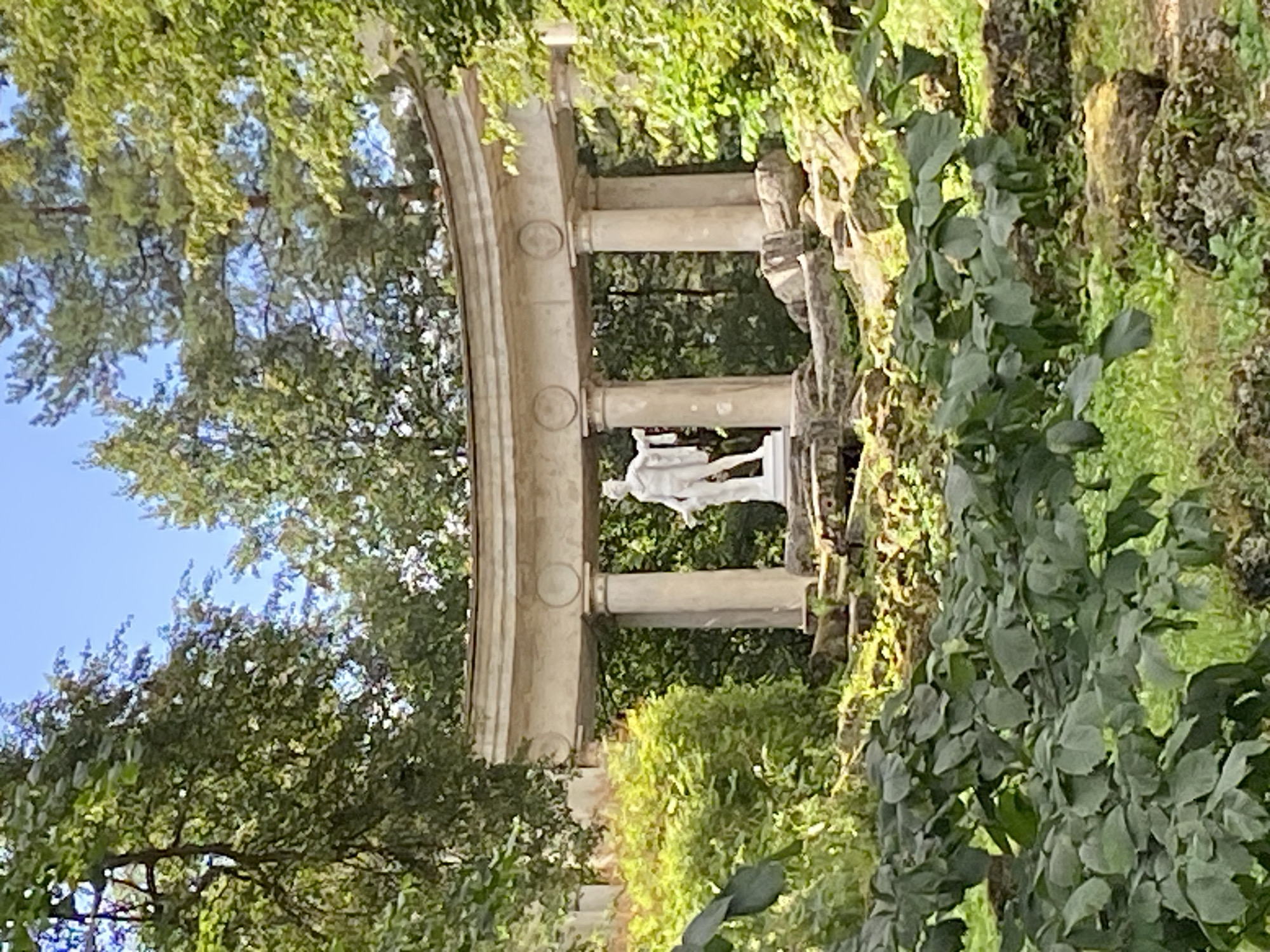 A white marble statue of a male figure stands in the center of the frame, positioned within a classical colonnade structure. The statue is located in Pavlovsk, Russia, amidst lush greenery and trees. The colonnade features a curved entablature supported by multiple columns. In the foreground, dense green foliage partially obscures the view of the statue and colonnade. The background is filled with the canopy of tall trees, with a bright blue sky visible through the leaves. The environment appears to be a park or garden setting, suggesting a historical or cultural landmark.