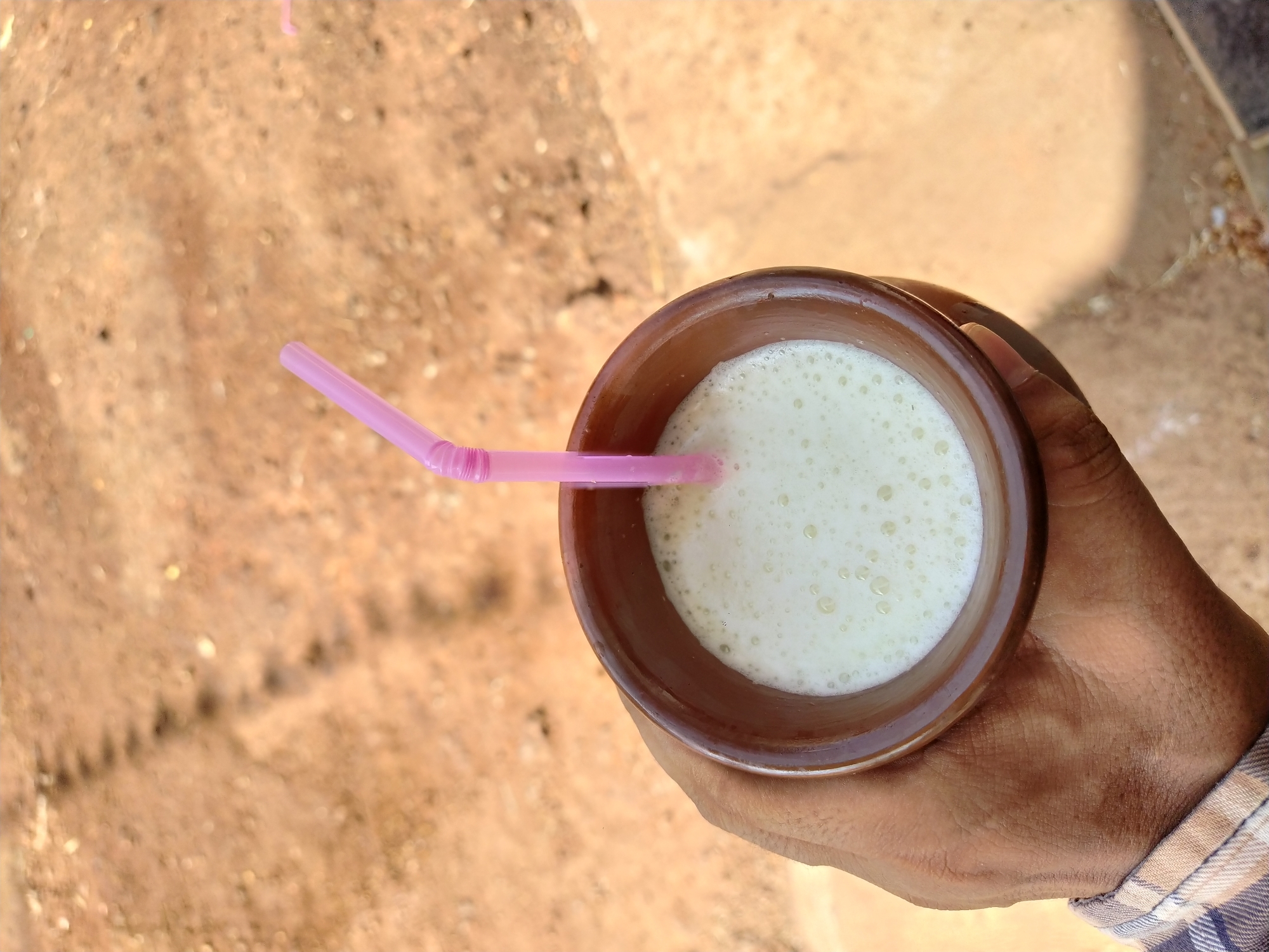 A top-down shot shows a person holding a traditional earthenware cup containing a light-colored, frothy liquid, likely a beverage. A pink plastic straw is inserted into the drink. The person's hand is visible, and the cuff of a patterned shirt or garment can be seen on their wrist. The background appears to be a rough, earthen surface. This scene may be located in Mapusa, India, based on cultural elements of the cup and potential local beverages.