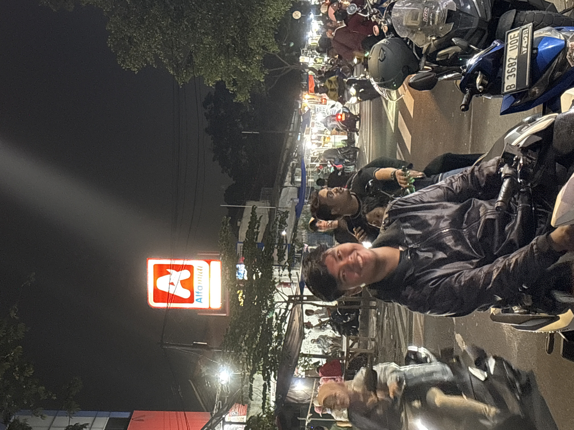 The image, taken at night, shows a bustling street scene in North Jakarta, Indonesia. Dominating the view is an illuminated "Alfamidi" convenience store sign. A crowded street market or food stalls are active. Several people are visible in the foreground, including a man on a motorcycle facing the camera and smiling. Another man appears to be drinking a bottle of beer in the middle-ground. Motorbikes are densely packed along the road, including one with the license plate "B 3682 UOE." Overall, the image depicts a lively evening scene in a busy urban environment.