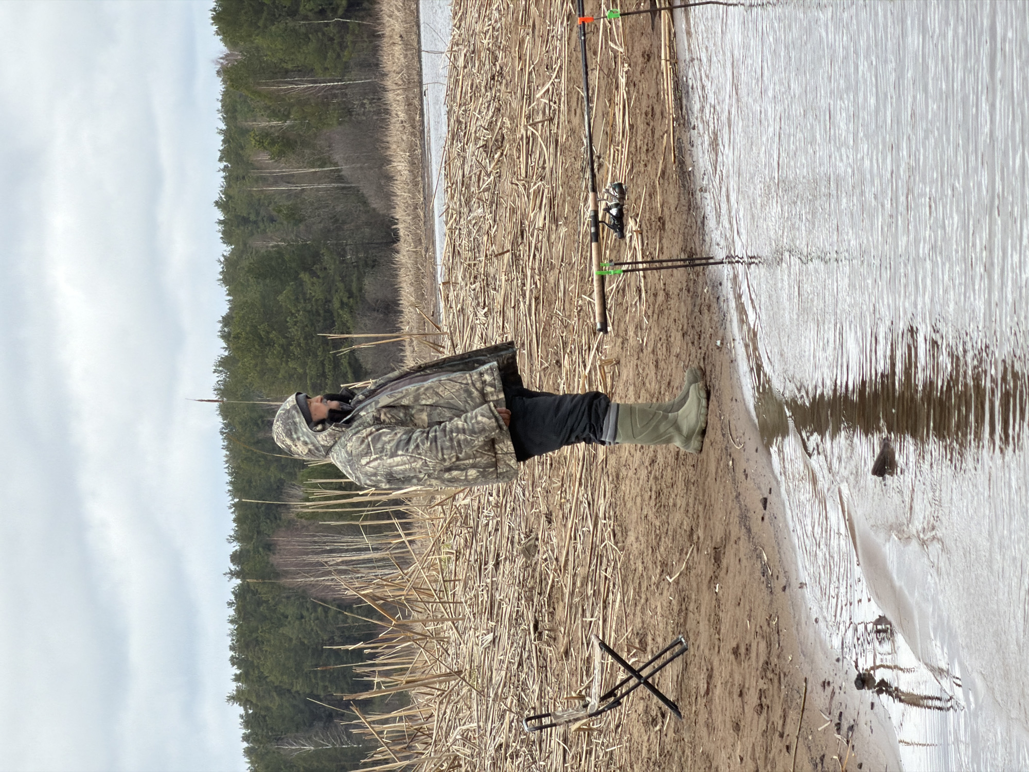 A person in camouflage clothing is fishing by a body of water. The person is wearing boots and standing on a sandy bank covered with dry reeds. Two fishing rods are set up nearby. A small folding chair is on the ground. In the background is a forest and cloudy sky. The location is likely in or near Chaykovsky, Russia, given the request.