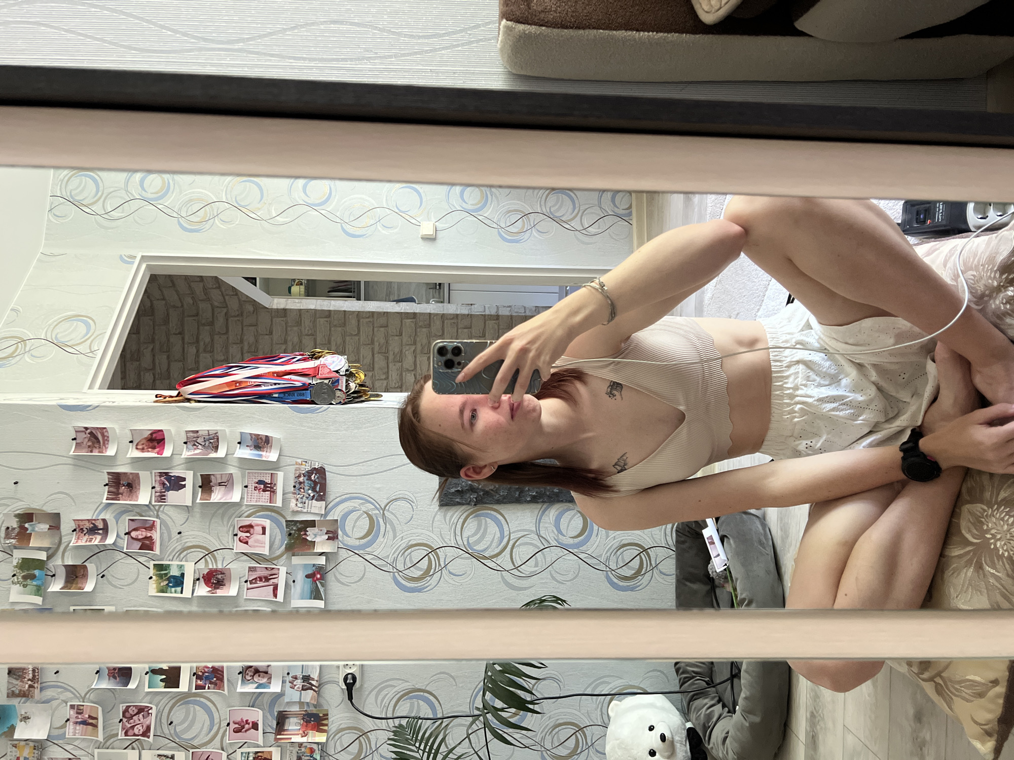 The image captures a young woman taking a selfie in a mirror within what appears to be a domestic setting. The background features wallpaper with a repeating circular pattern, and a wall adorned with numerous photographs, along with a shelf displaying medals and ribbons. A doorway leads to a room with brick-like walls. In the foreground, there's a potted plant and a stuffed animal visible. The woman is wearing a light-colored top and shorts, and a watch is visible on her wrist. Based on the metadata and clues like the Russian medals, this image is likely taken in Chaikovsky, Russia.