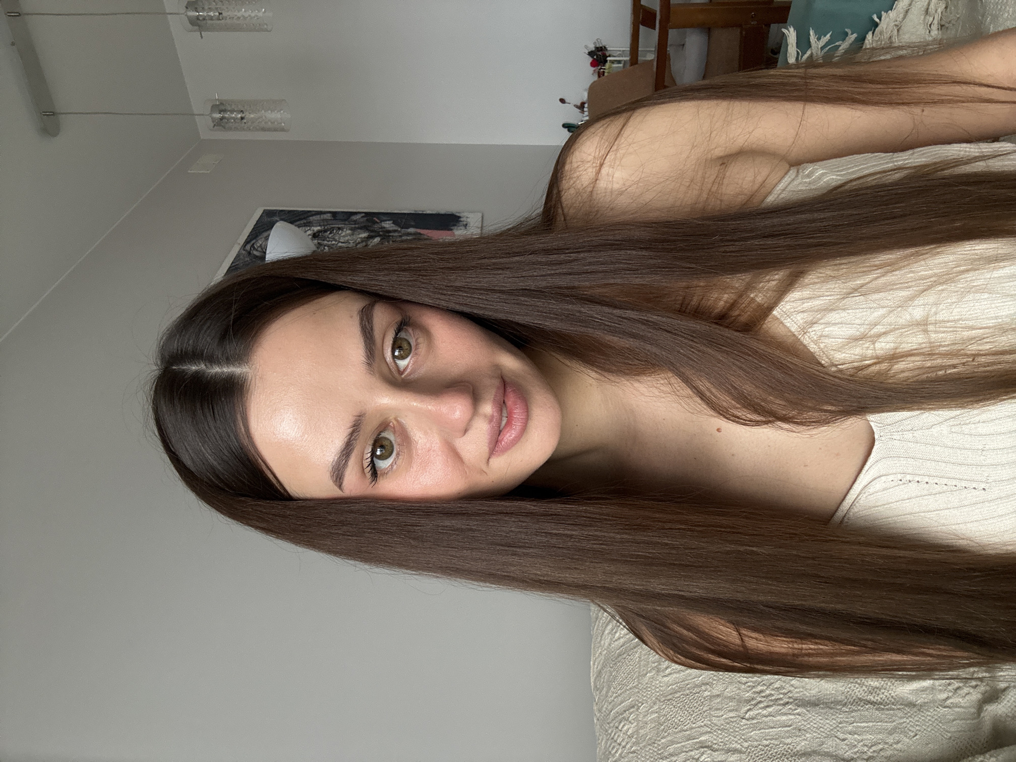 A young woman with long, straight brown hair is lying down indoors, possibly on a bed or sofa. She's wearing a light-colored top. Her face is centered in the frame, showing clear skin, hazel eyes, and dark eyebrows. The background includes a neutral-colored wall, with a decorative lamp and an artwork partially visible. A wooden furniture piece is slightly out of focus in the background, but the image provides no information to establish the specific location within Krasnoyarsk, Russia.