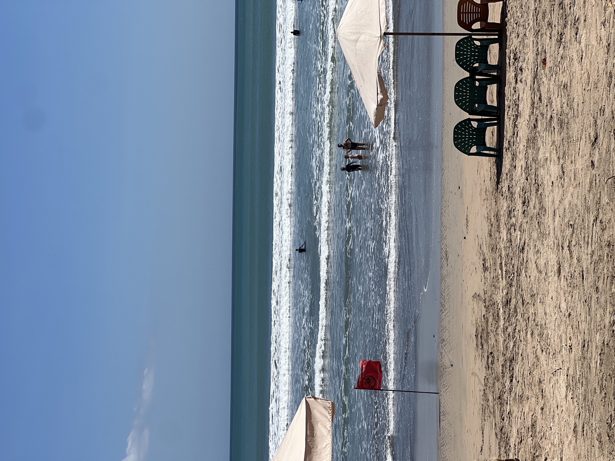 This is a medium shot of a beach in Kuta, Indonesia, on a bright, sunny day with a clear blue sky. The foreground shows a sandy beach with three green plastic chairs and one brown plastic chair arranged in a row under a white beach umbrella. Further back, a red flag is planted in the sand near the water's edge, indicating "swimming prohibited" with a graphic of a person being pulled underwater. Beyond the flag, the ocean stretches out with moderate waves breaking. Several people are visible in the water, including a lone surfer further out and a group of three people wading near the shore. Another person is standing in the water on the right side of the frame. A second white beach umbrella is partially visible on the left side of the image. The overall scene depicts a beach with a warning for swimmers, suggesting potentially strong currents or other hazards.