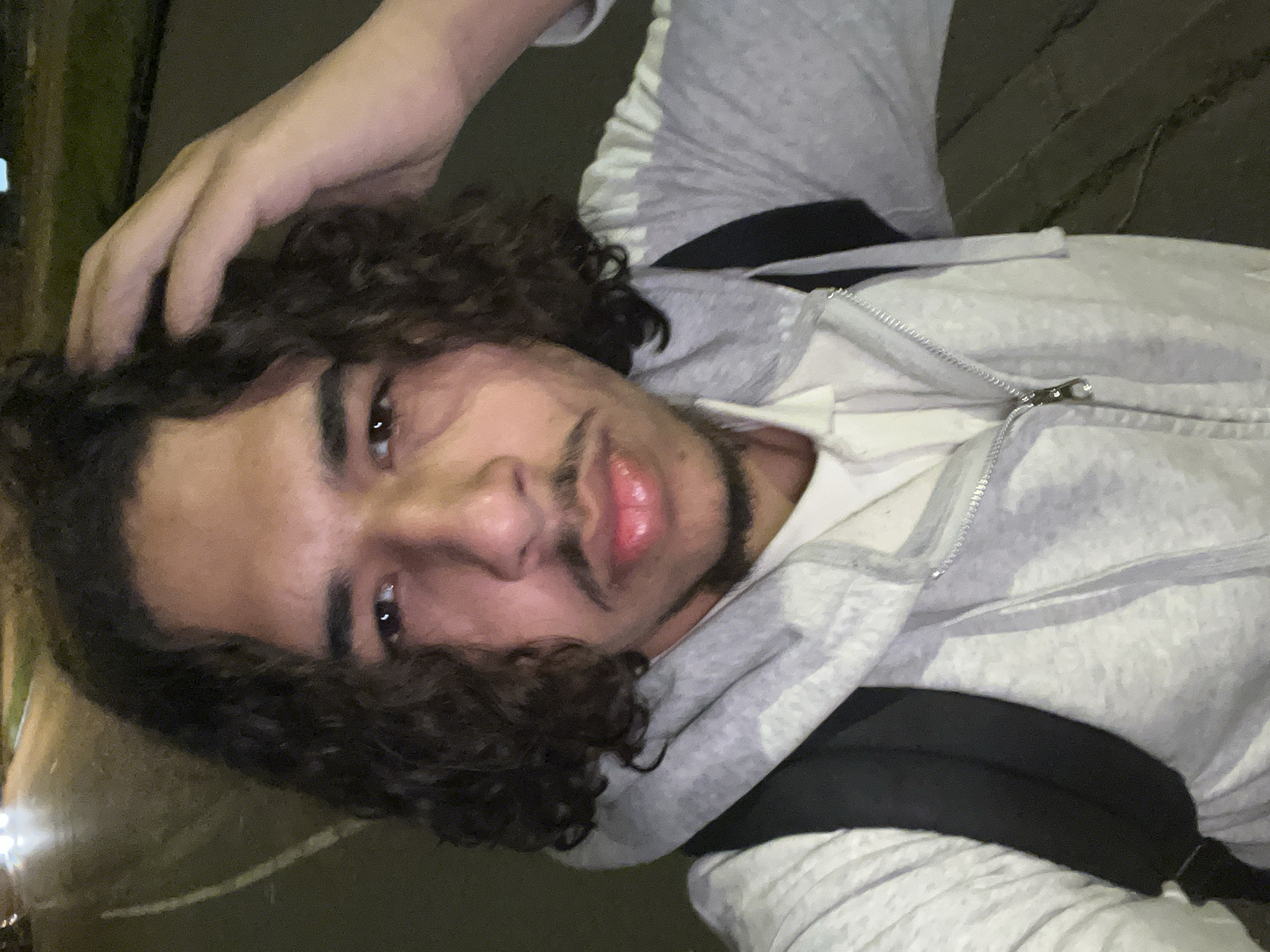 A young man with curly dark hair and a slight mustache poses for a close-up selfie at night. He is wearing a light grey hooded sweatshirt partially zipped, revealing a white t-shirt underneath. He has a black strap, presumably from a backpack, across his chest. His right hand is raised and slightly touching his hair. The setting is dark, possibly outdoors, with a blurry, indistinct green and white light source in the background. The location is unspecified and the city of Hoenheim, France is not discernible from the image.