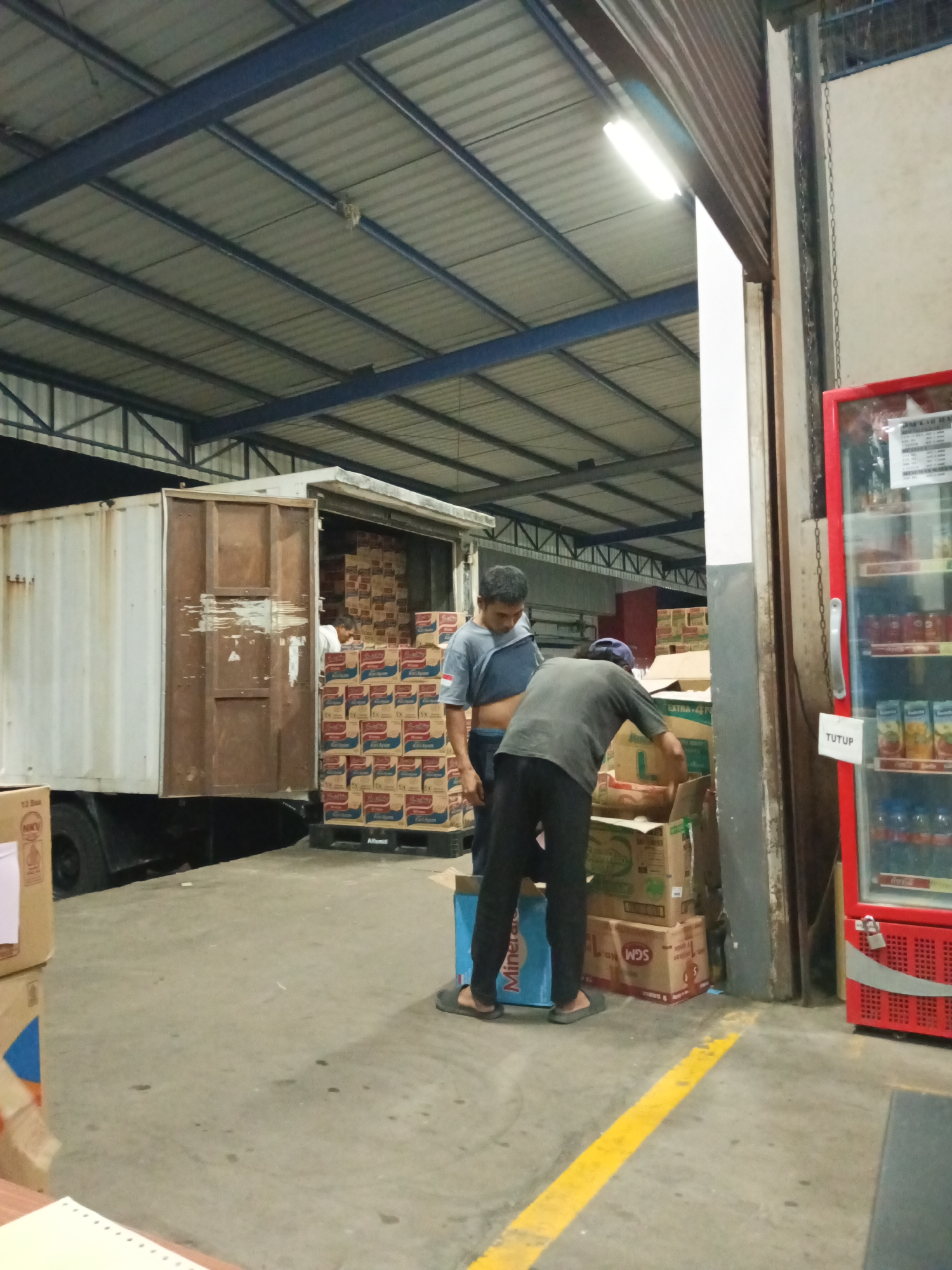 The image depicts a warehouse environment in Makassar, Indonesia, with two men engaged in loading or unloading goods from a truck. The warehouse features a corrugated metal roof supported by dark blue steel beams, and fluorescent lighting fixtures are visible. In the foreground, a red refrigerator with a glass door displays various beverage bottles and appears to be locked. A "TUTUP" (close) sign is affixed to its door. The ground is a concrete floor marked with a yellow line. The truck, partially loaded with boxes, is positioned to the left of the frame. The men are interacting with stacks of boxes, some of which are labeled with brands. The overall scene suggests a commercial or industrial setting involved in logistics and distribution.
