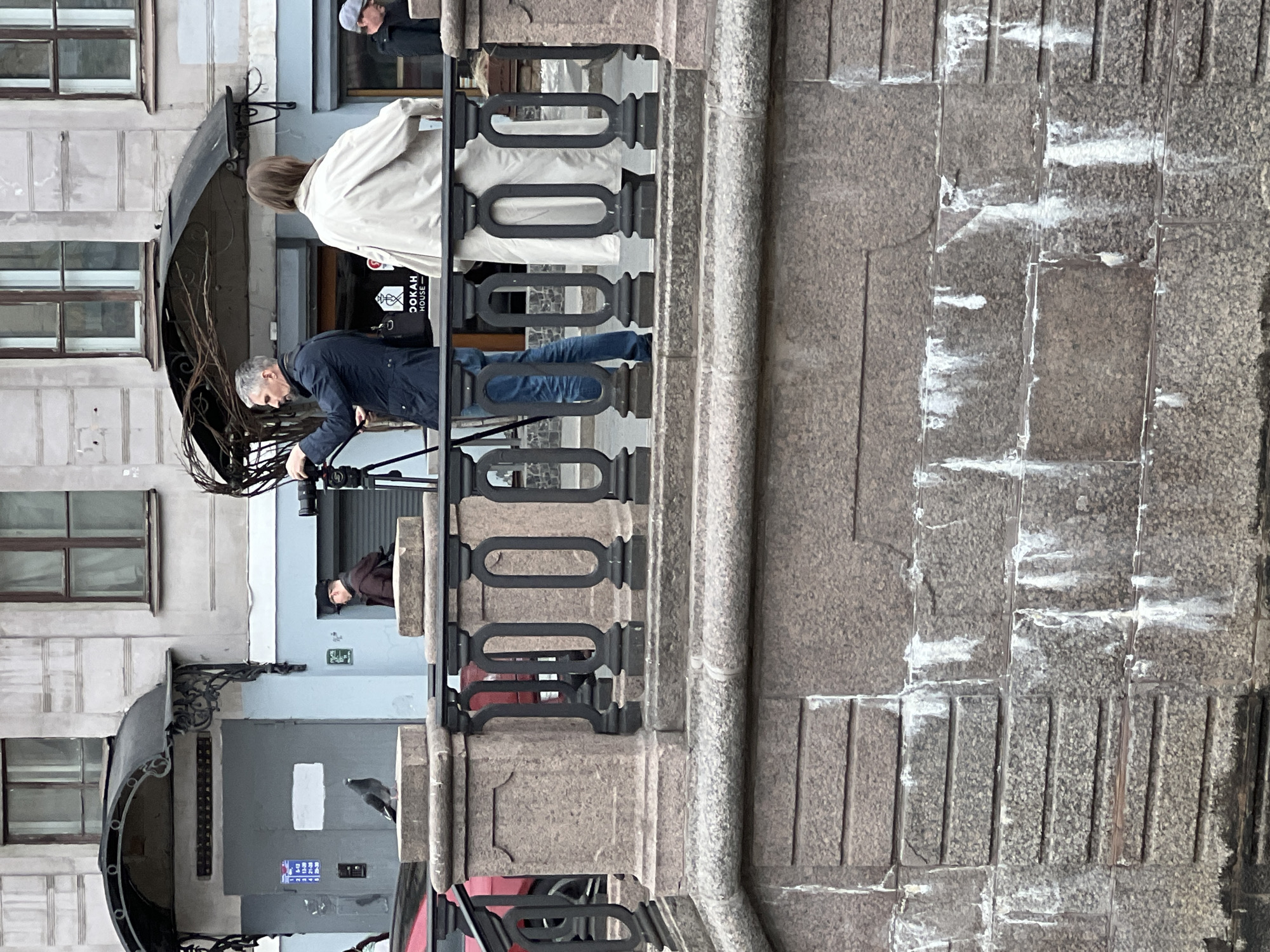 An outdoor scene in Saint Petersburg, Russia, captured during the daytime, depicts a photography or filming session in an urban setting.
In the foreground, a coarse, grey-brown stone surface with pronounced white streaks and vertical grooves takes up the right side of the image, suggesting a pavement, wall, or bridge structure. A black, ornate metal railing or balustrade runs horizontally across the middle ground, separating the stone surface from the background elements.
Behind the railing, to the left, two individuals are prominent. A man, wearing a dark blue jacket and light grey hair, is crouched and focused on operating a camera mounted on a black tripod. He appears to be setting up or adjusting the camera. Standing slightly behind and to his right is a woman with light brown hair, wearing a light-colored, possibly cream or beige, trench coat or jacket. She is looking in the direction the camera is pointing, seemingly observing the scene or waiting for instructions.
Further back, behind the man and woman, are parts of building facades. To the left, a light-colored building with dark-framed windows is visible. Below it, an ornate dark awning extends over a doorway. On a black sign above this doorway, the text "DOOKAH HOUSE" is clearly visible in white letters, with a circular emblem to its left. To the right of this, another light blue building facade is partially seen, with a grey door and a small blue sign with white pictograms (likely indicating a restroom or accessibility). Another person in a dark jacket and a cap is partially visible through a window opening in the upper right background. A red object is partially visible below the railing in the lower right.
The overall impression is of professionals engaged in media production in a city environment.