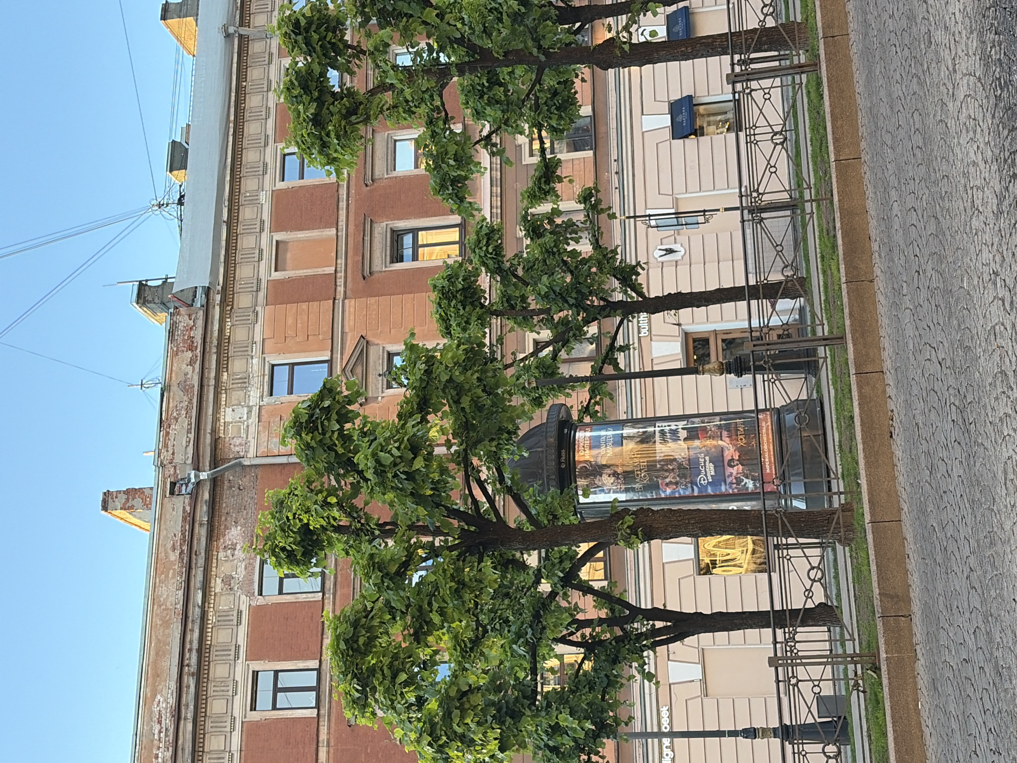 The image shows a street scene in Saint Petersburg, Russia, with a multi-story building in the background. The building has red brick and ornate detailing. In the foreground, there are several trees with green foliage, a street lamp, and a metal fence. A cobblestone street is visible at the bottom of the image. There are shops on the ground floor of the building, with awnings and signage. A large cylindrical display with posters is situated between two trees. The sky is clear and blue.