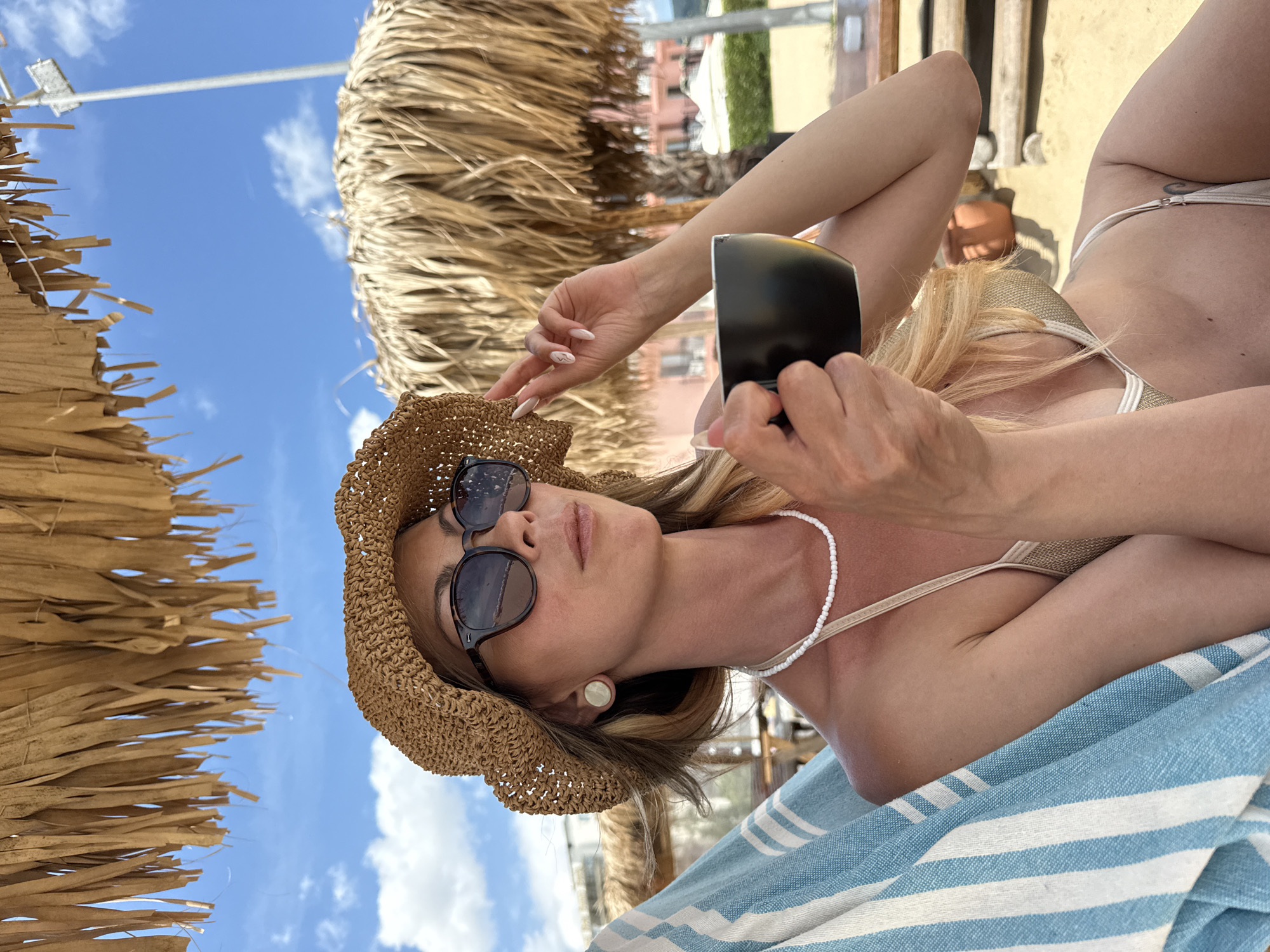 A woman with blonde hair wearing a straw hat and sunglasses sits in a beach chair under a thatched umbrella. She is holding a black mug of coffee in her right hand and her left hand is resting on her hat. She is wearing a gold bikini and a white beaded necklace. A blue and white striped towel is draped over the chair. In the background, there are more thatched umbrellas, a beach, and buildings. The sky is blue with some clouds. The location is Nesebar, Bulgaria.
