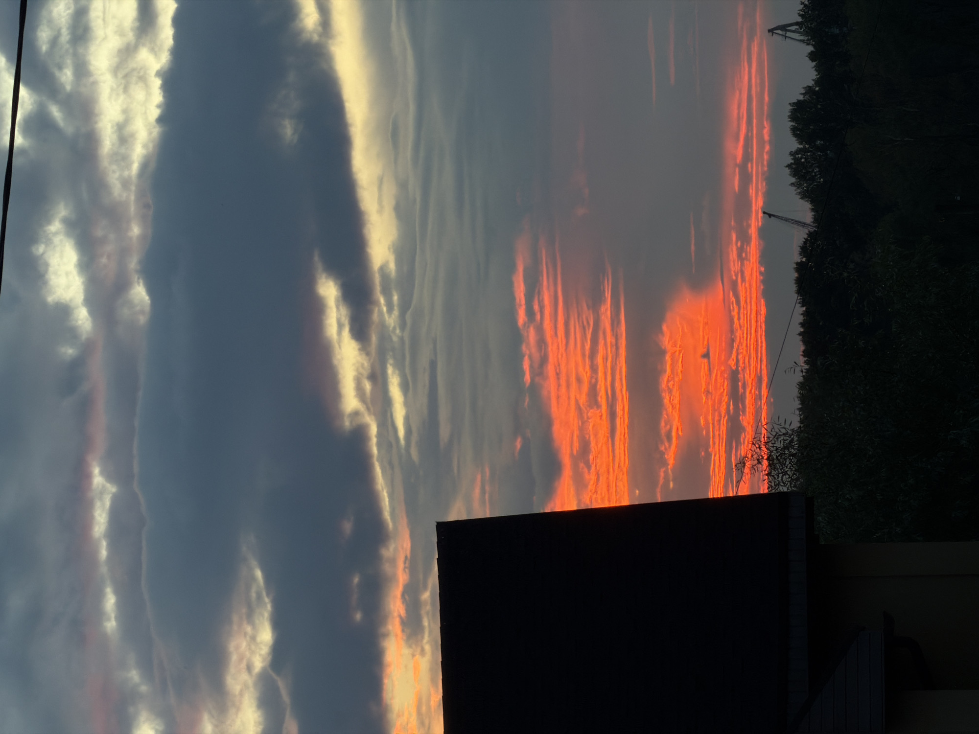 The image depicts a dramatic sunset over Chaikovsky, Russia. The sky is a canvas of vibrant colors, with fiery orange clouds illuminated by the setting sun, contrasting with darker, heavy grey clouds above. In the foreground, the dark silhouette of a building's roof juts into the frame from the left, casting a shadow. Below the horizon, a dense line of dark green trees forms the immediate landscape. In the distance, two industrial cranes are visible against the colorful sky, hinting at the urban or industrial presence within the otherwise natural scene. A thin black wire is visible stretching across the upper portion of the image, suggesting overhead power lines. The overall impression is one of a stark and visually striking end to the day.