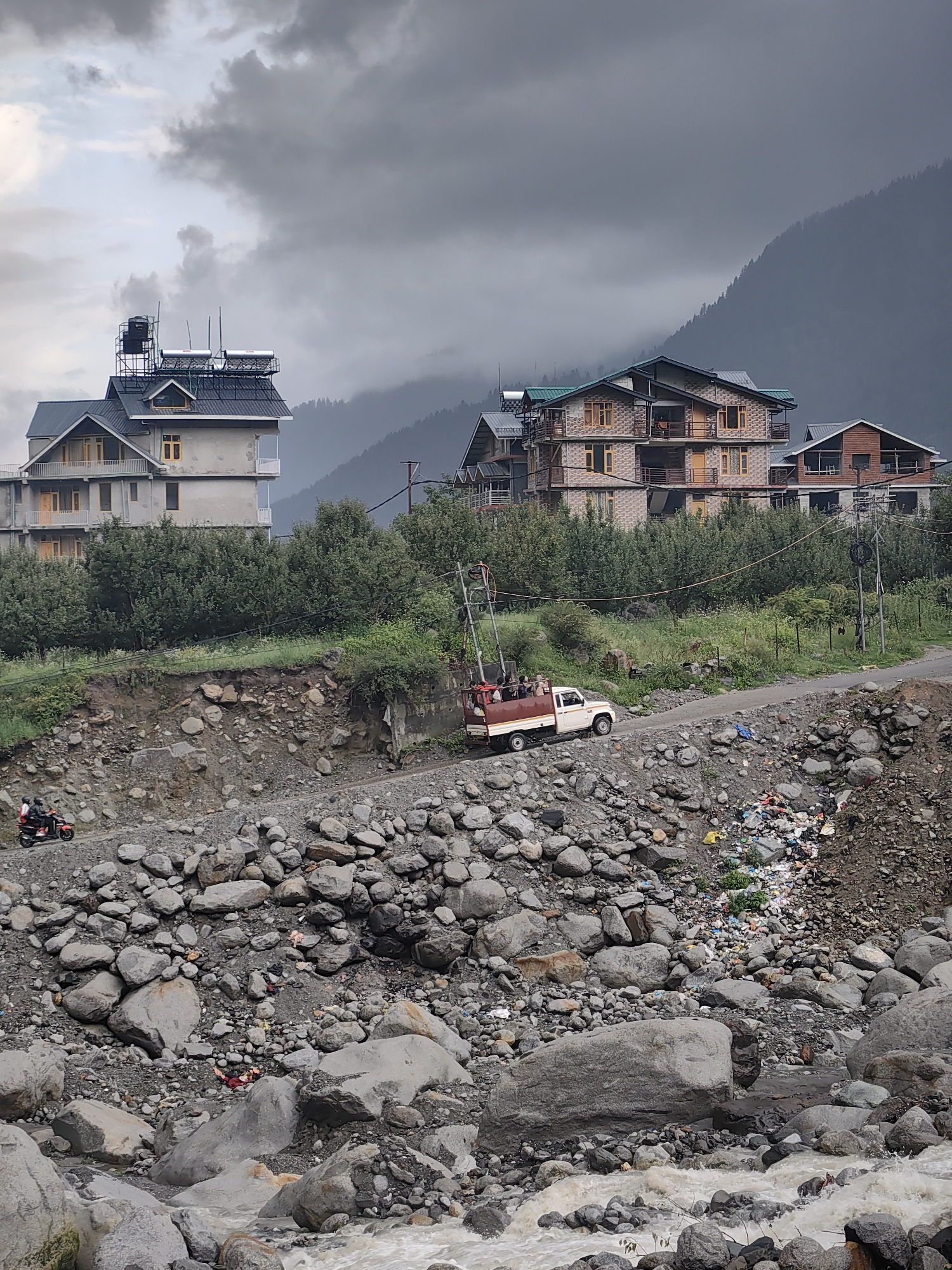 The image captures a rural scene in Palchan, India, with a winding dirt road leading up a rocky embankment. On the left, two individuals ride a red scooter uphill. Further up the road, a white pickup truck carries several passengers. A cluster of colorful plastic waste is visible on the embankment. In the background, a large, multi-story building with a dark roof and solar panels is situated on a hillside, with a misty, tree-covered mountain rising behind it. Several other structures, likely houses, are also visible on the slopes. The environment is characterized by rough terrain, large rocks, dense green foliage, and a cloudy, overcast sky.