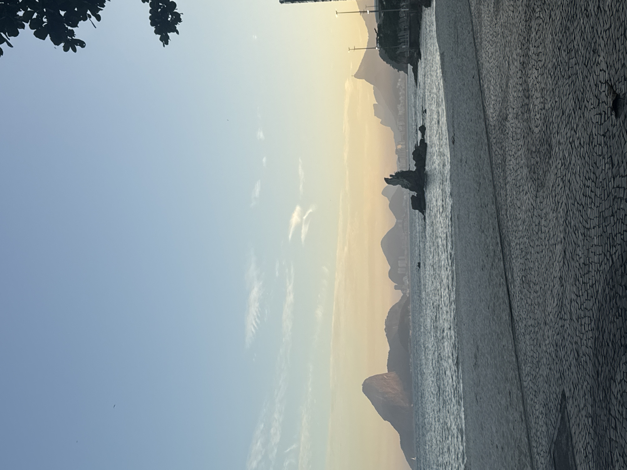 The image shows a coastal scene with a clear sky and calm water. In the foreground, there is a paved walkway with a pattern of dark stones, leading to a sandy beach. Beyond the beach, the ocean stretches out, with gentle waves. In the distance, silhouetted against a soft, hazy sky with hues of yellow and orange, are several prominent mountains and a city skyline. One of the mountains has a distinctive, jagged peak, and another, further to the right, appears to have a structure on its summit. The overall atmosphere is serene, suggesting a tranquil moment at dawn or dusk. This location is likely the Praia de Icaraí in Niterói, Brazil, known for its iconic view of the Rio de Janeiro skyline across Guanabara Bay, featuring mountains like Pão de Açúcar and Corcovado.