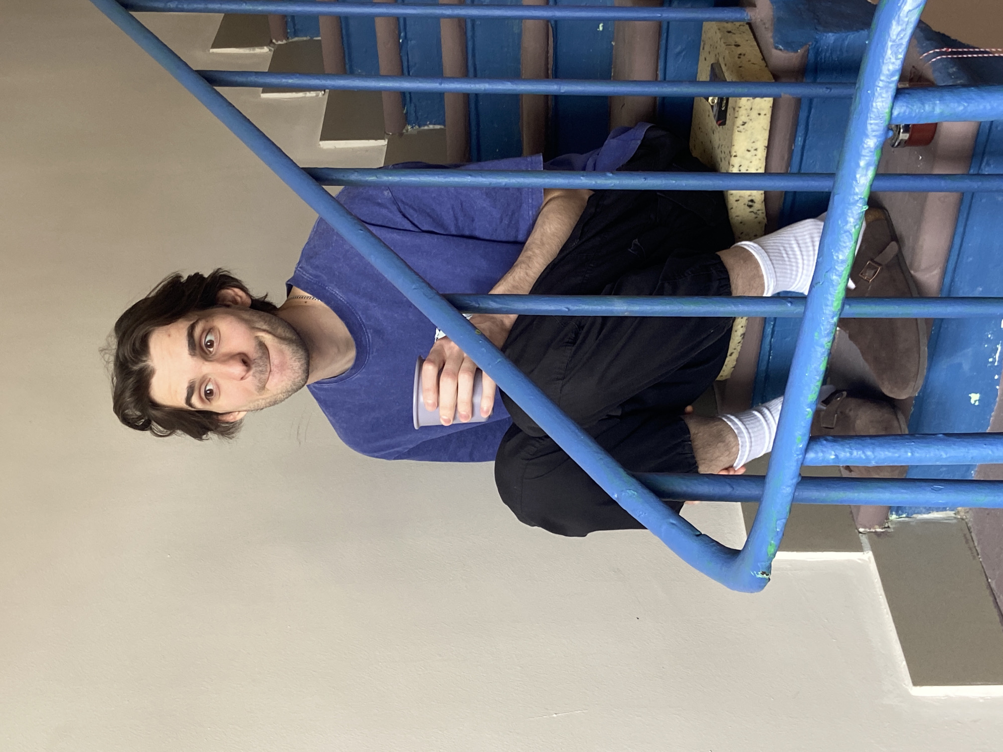 The image, rotated counter-clockwise, shows a young man with dark hair and stubble, sitting on a staircase in what appears to be an indoor or covered public building, possibly in Moscow, Russia. He is positioned behind a prominent blue metal railing, which takes up a significant portion of the foreground and midground, running diagonally across the frame.
The man is wearing a casual blue t-shirt and black pants, with white socks visible above brown shoes or boots. He is holding a light-colored cup in his left hand. He looks directly at the camera with wide, brown eyes and a slight, curious smirk. His body is turned slightly to his right, and his legs are bent, indicating a squatting or seated posture on the stairs, partially obscured by the railing.
The background consists of light-colored walls and the steps of a concrete staircase. Behind the man, a section of yellow, speckled foam material is visible, possibly part of a makeshift cushion or an exposed insulation layer. The blue metal railing, which has some chipped paint revealing glimpses of green and bare metal, is composed of vertical and horizontal bars, characteristic of utilitarian staircases found in older apartment buildings or public structures in post-Soviet areas. The overall lighting suggests an indoor setting during the daytime. There is no visible text in the image.