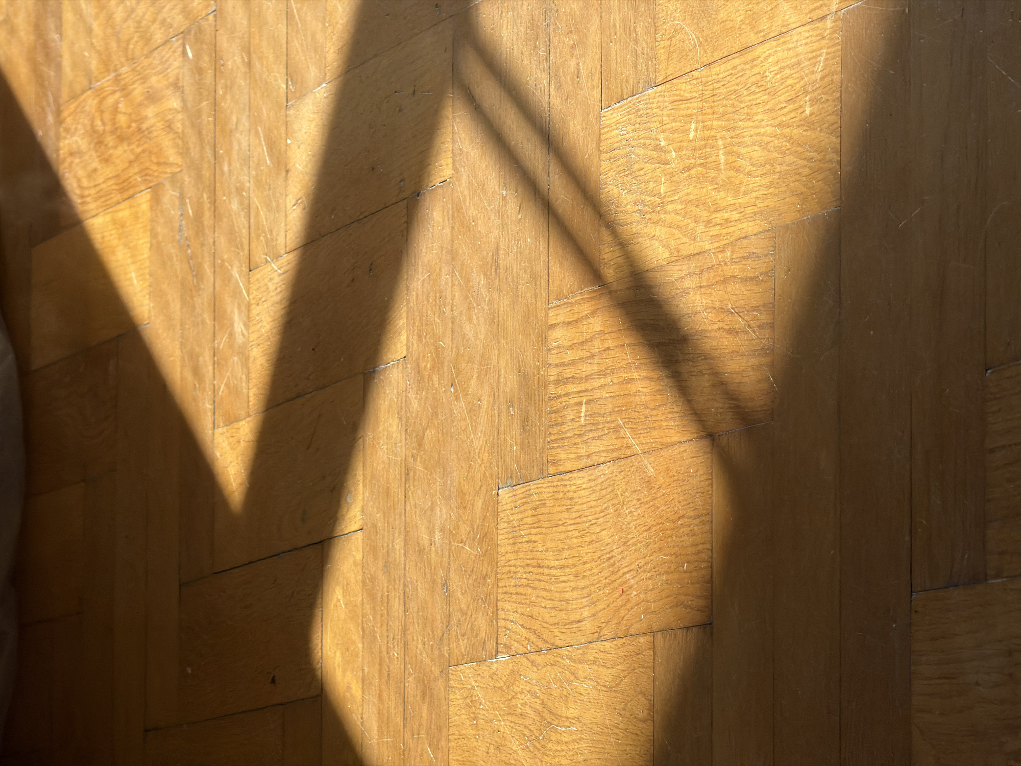 The media file shows a close-up, overhead view of a wooden floor with a parquet pattern. Sunlight streams in from the left, casting distinct shadows across the floor. The wood appears to be a light to medium brown, with visible grain patterns and some scratches or scuff marks, indicating wear. The parquet arrangement consists of rectangular wooden blocks laid in a herringbone or chevron pattern, with some blocks oriented at an angle to others.
The shadows are sharp-edged, suggesting a bright light source, likely the sun, positioned at an angle. One large, triangular shadow dominates the left side of the frame, with thinner, parallel shadows extending diagonally across the mid-right portion. These shadows suggest the presence of an object or structure outside the frame, such as blinds, a window frame, or furniture, casting its silhouette onto the floor.
The lighting and the stark shadows suggest it is daytime, likely late morning or early afternoon when the sun is high enough to create such strong contrasts. The overall impression is one of a quiet, indoor space. There are no people or distinct objects clearly visible, with the focus solely on the texture of the wood and the play of light and shadow. The scene is neutral and devoid of overt emotional cues, but the strong sunlight might evoke a sense of warmth or a passing moment. There is no visible text in the image.