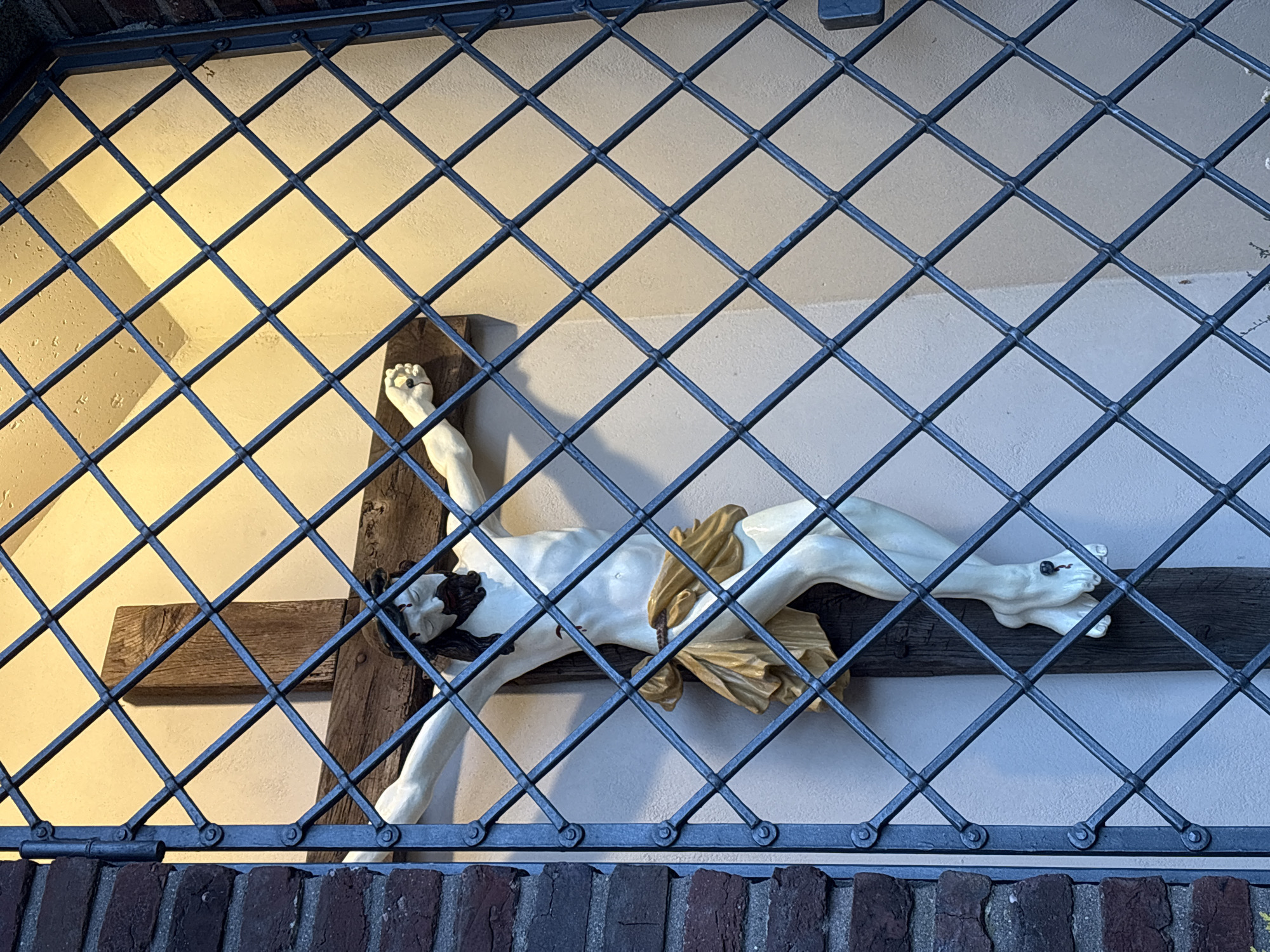 A statue of Jesus Christ on a cross, viewed through a dark metal diamond-shaped cage. The statue is white with brown hair and a golden cloth covering his lower body. The cross is made of wood. The cage is mounted on a brick structure. The background consists of two shades of off-white wall. The location is in Mönchengladbach, Germany.