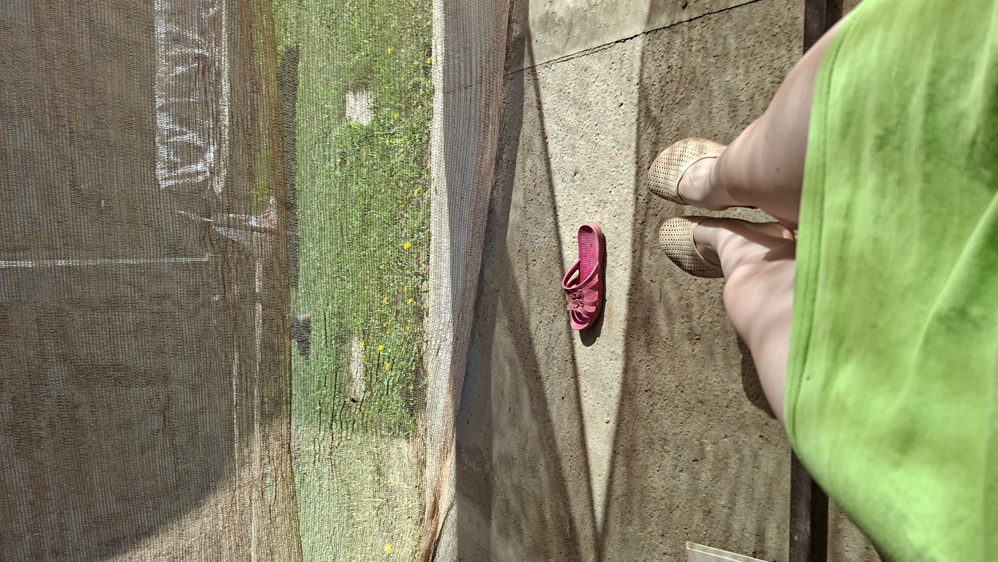 The image shows a person's legs and feet wearing beige woven flats standing on a concrete surface with shadows. The person is wearing a bright green dress or fabric. A single pink child's sandal lies on the concrete. To the left, there is a mesh screen obscuring a view of grass and vegetation, possibly a garden. Part of a building or structure can be seen through the screen. The photo appears to be taken outdoors on a sunny day. It is difficult to confirm the location is in Большой Букор, Russia, based solely on the visual information.