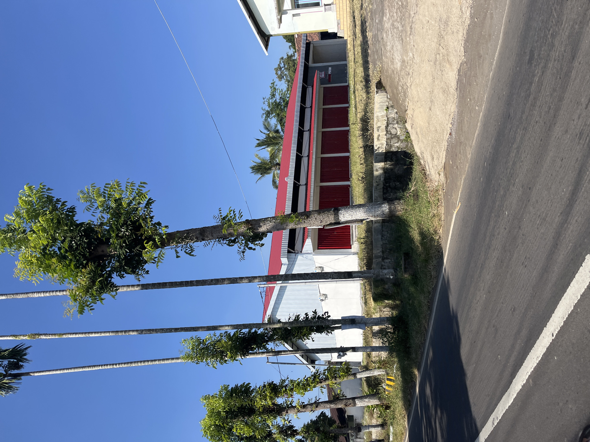 This is a photograph of a building with a red roof and white siding located in Purworejo, Indonesia. The building appears to be a commercial establishment, possibly a warehouse or a shop, with several red doors. In front of the building, there are several tall trees and a paved road. The sky is clear and blue, suggesting a sunny day. The overall scene depicts a typical street view in a rural or semi-urban area of Indonesia.