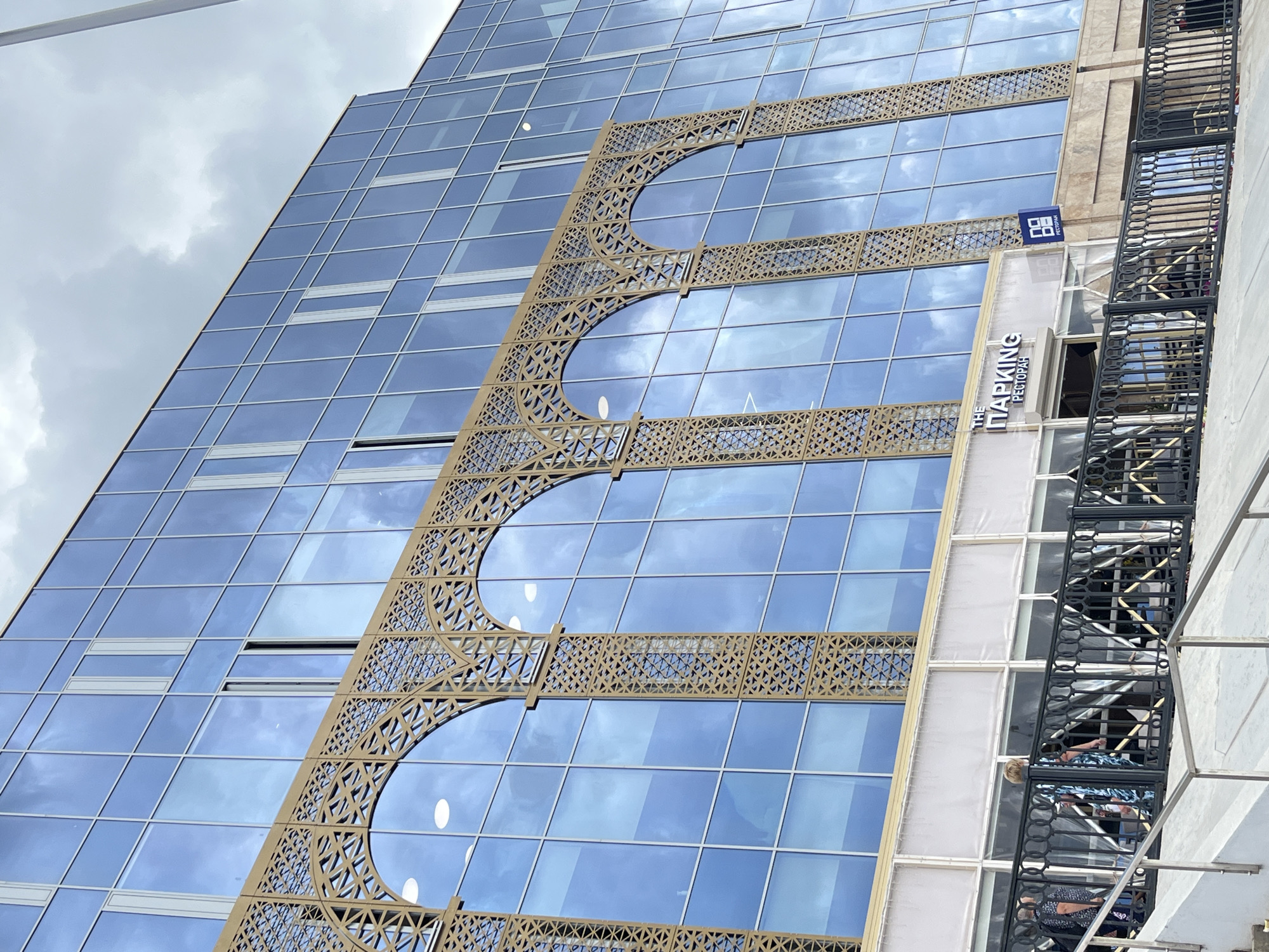 The image shows a modern building with a glass facade, featuring a decorative gold screen with arched patterns. Below the screen, a white awning covers a section of the building, where a sign reads "THE PARKING РЕСТОРАН". A blue sign with a square logo and "РЕСТОРАН" is also visible. In front of this is a black metal railing, with people walking behind it. The building appears to be located in the city of Perm, Russia, as indicated by the Cyrillic text on the signs. The sky above is cloudy, with reflections of the sky and clouds visible in the glass of the building. A set of concrete stairs leads down to the foreground of the image.