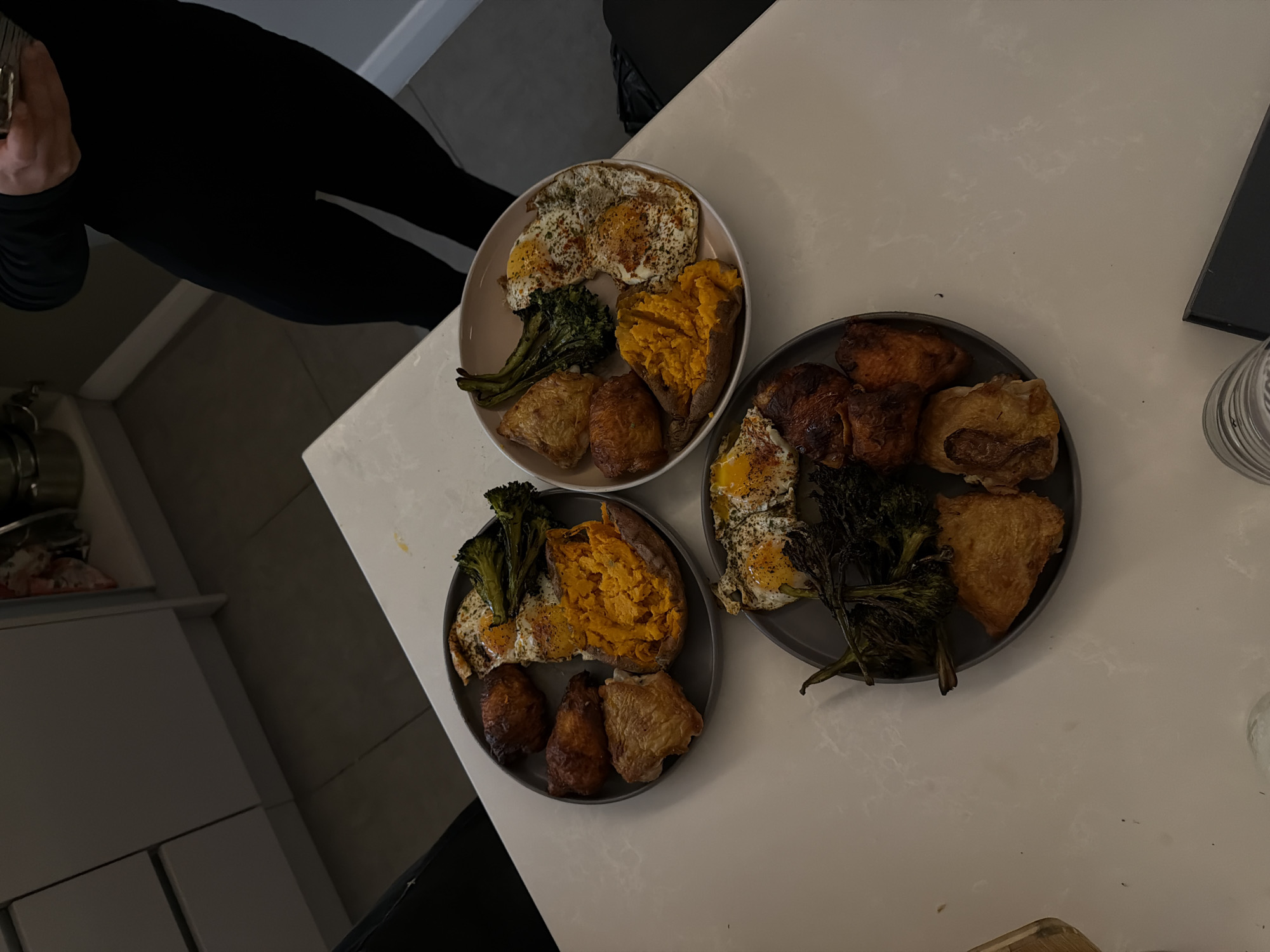 The image presents an overhead view of a kitchen or dining area, featuring three plates laden with food on a light-colored countertop. A person is partially visible on the far left, clad in black long-sleeved attire and pants, with their hands holding an object not entirely visible, possibly cutlery or a phone.
Each of the three plates holds an identical meal. The main components include two fried eggs, seasoned with what appears to be paprika or red pepper flakes and black pepper. Alongside the eggs, there is a large baked sweet potato, its vibrant orange flesh evident, suggesting it has been softened or slightly mashed. Roasted green vegetables, likely broccoli or broccolini, some with a slightly charred appearance, are also present on each plate. Completing the meal are several pieces of roasted chicken, possibly thighs or drumsticks, distinguished by their crispy, golden-brown skin.
In the background on the left, kitchen cabinets or drawers are visible, along with a metallic pot or pan on a shelf. On the right side of the countertop, a clear glass or cup can be seen, and a dark, flat object, possibly a laptop or a book, rests near the edge. The indoor lighting suggests the photograph was taken during the evening or night. The scene captures a prepared meal, possibly indicating dinner for multiple individuals or meal prepping. There is no visible text in the image.