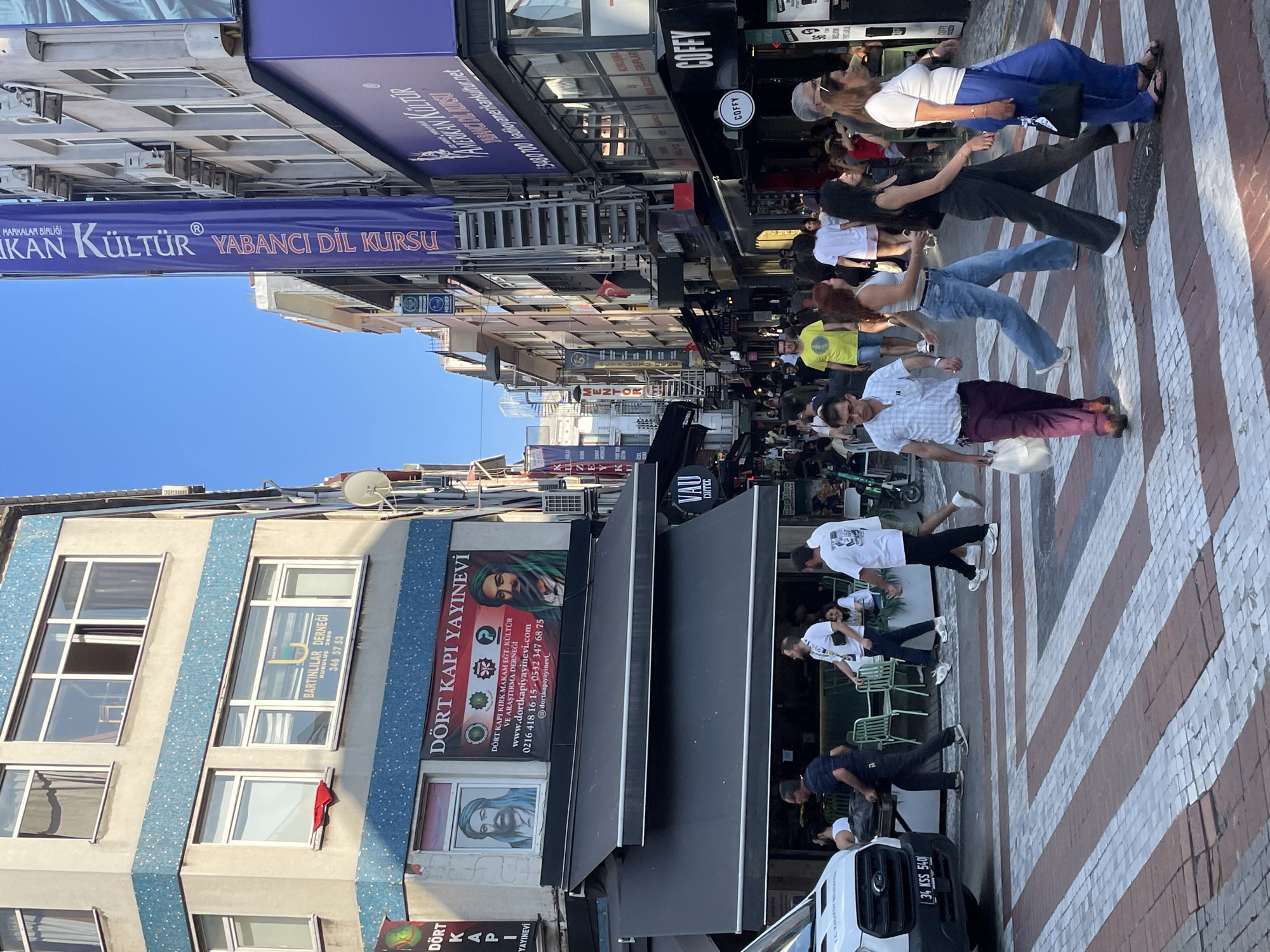 The image depicts a busy street scene in Kadıköy, Turkey, likely on a sunny day. In the foreground, a pedestrian crossing with white and reddish-brown stripes is visible. Several people are crossing the street. One man in a light blue checkered shirt and maroon pants carries a white plastic bag. A woman in a grey tank top and blue jeans walks beside him. Further ahead, a woman in a black tank top and grey pants walks with a black handbag. Another woman in a white top and blue pants is also crossing.
The background shows a street lined with buildings housing various businesses. To the left, a white van with the license plate "34 KSS 540" is parked. Above the van, a building has signs for "DÖRT KAPI YAYINEVI," which appears to be a publishing house. Further up this building, there are windows with posters and advertisements.
On the right side of the street, a large blue banner with white text reads "AMERİKAN KÜLTÜR YABANCI DİL KURSU," indicating an American Culture language school. Below this banner, there are shopfronts, including one with the signage "COFFY." People are seen sitting at tables outside some establishments. The architecture of the buildings suggests a commercial and residential area. The overall impression is a lively urban environment with commercial activity and pedestrian movement.