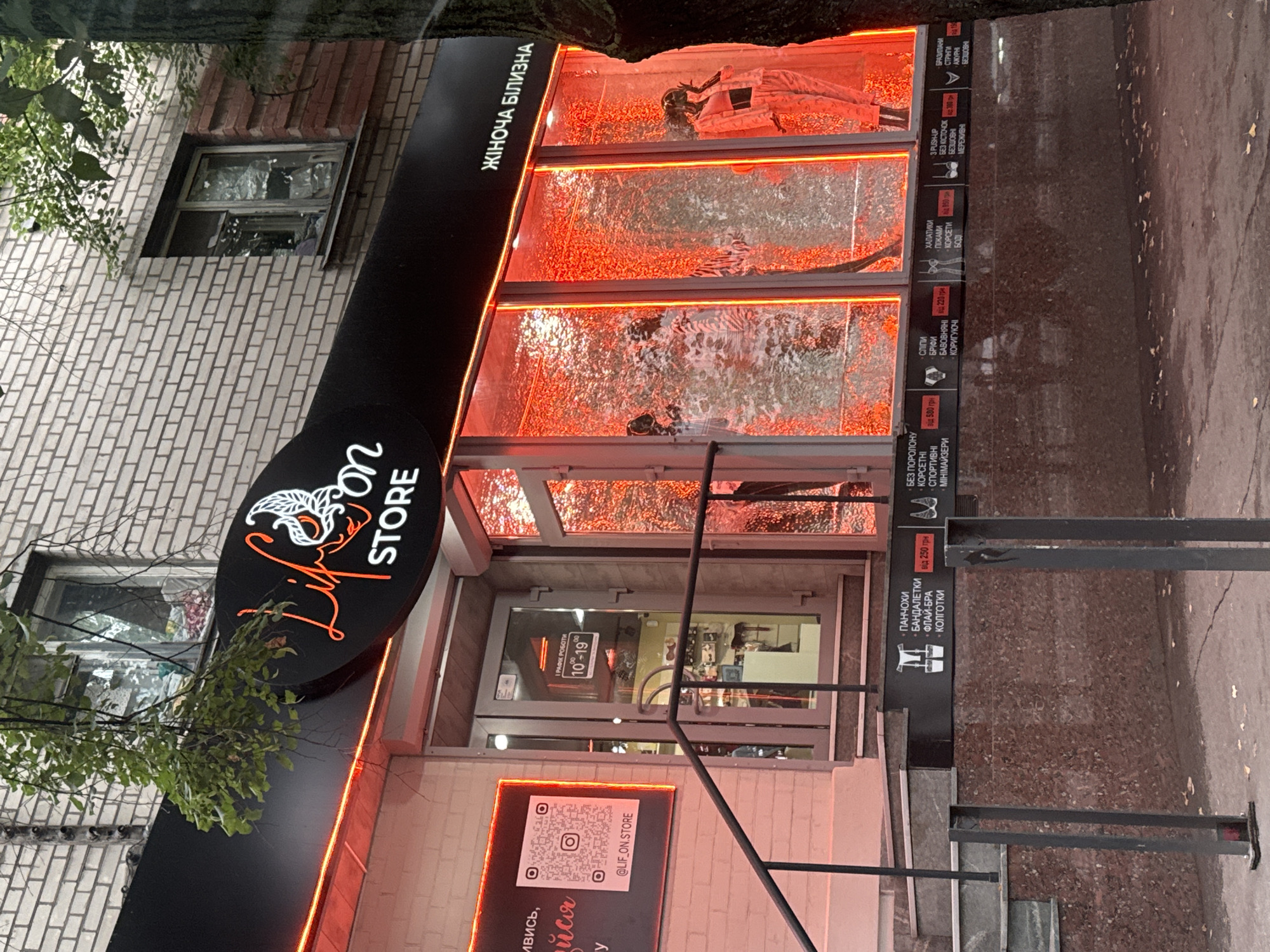 The image shows the storefront of a store named "Lif Store" located in the city of Dnipro, Ukraine. The store has a black awning with a neon red border and a circular logo featuring the store name and a stylized bird. The windows are illuminated with red lighting and feature mannequins dressed in what appears to be loungewear or sleepwear. A sign above the window on the right reads "ЖІНОЧА БІЛІЗНА," which translates to "Women's Lingerie." A QR code with the handle "@LIF_ON_STORE" is displayed on the left side of the building. Below the awning, a dark band displays various categories of women's undergarments with prices in Ukrainian Hryvnia. The store entrance is a glass door with a sign indicating operating hours from 10:00 to 19:00. The building is constructed of brick, and trees with green foliage are visible around the storefront.