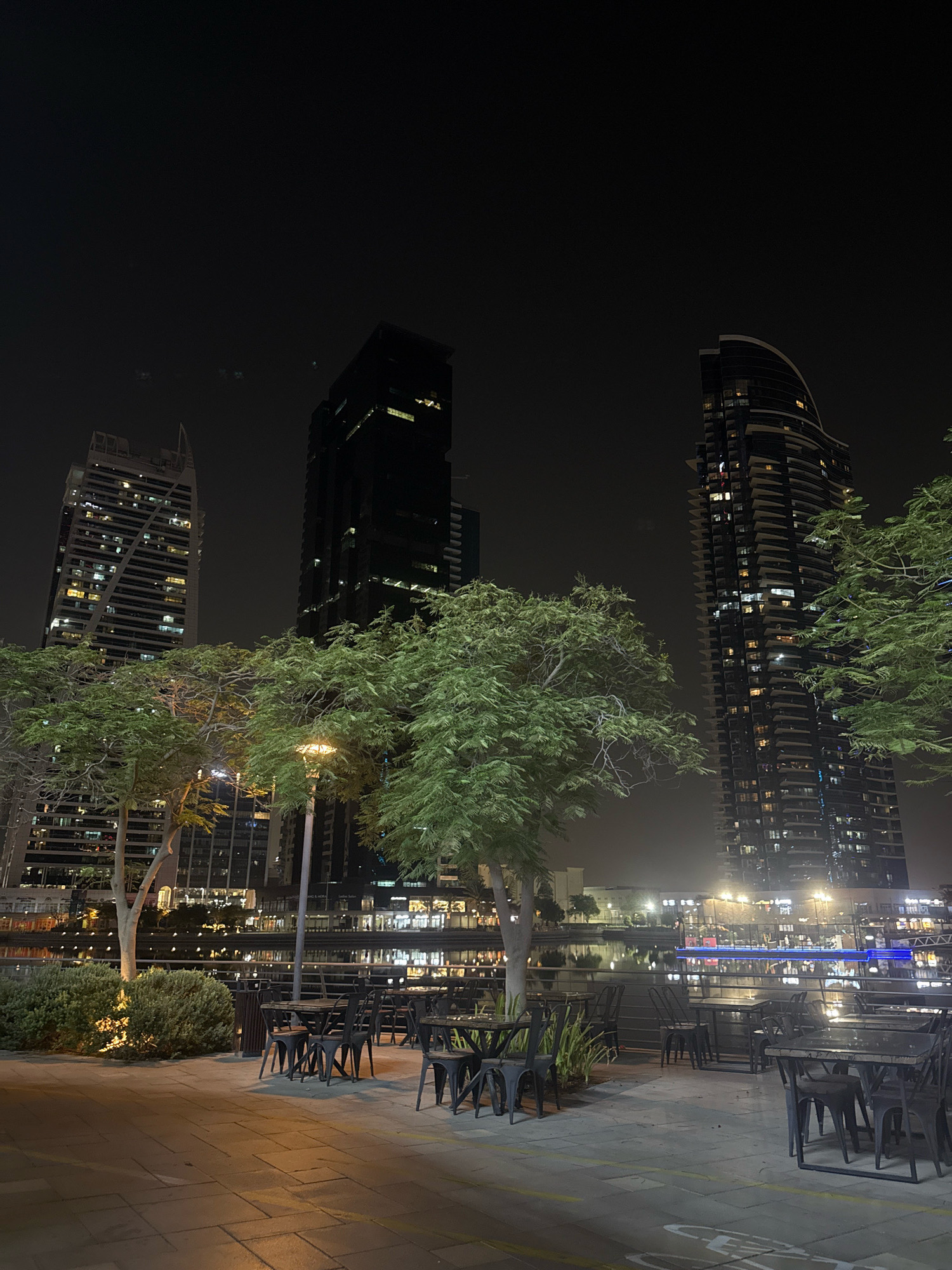 A night view of a waterfront dining area in Dubai Marina, United Arab Emirates. In the foreground, an empty outdoor seating area with dark metal tables and chairs is set up on a paved walkway. Beyond the seating area, lush green trees and bushes are illuminated by ambient lighting. Across the water, a strip of buildings with lights on display lines the waterfront, with several modern skyscrapers towering in the background. The sky is dark, indicating nighttime.