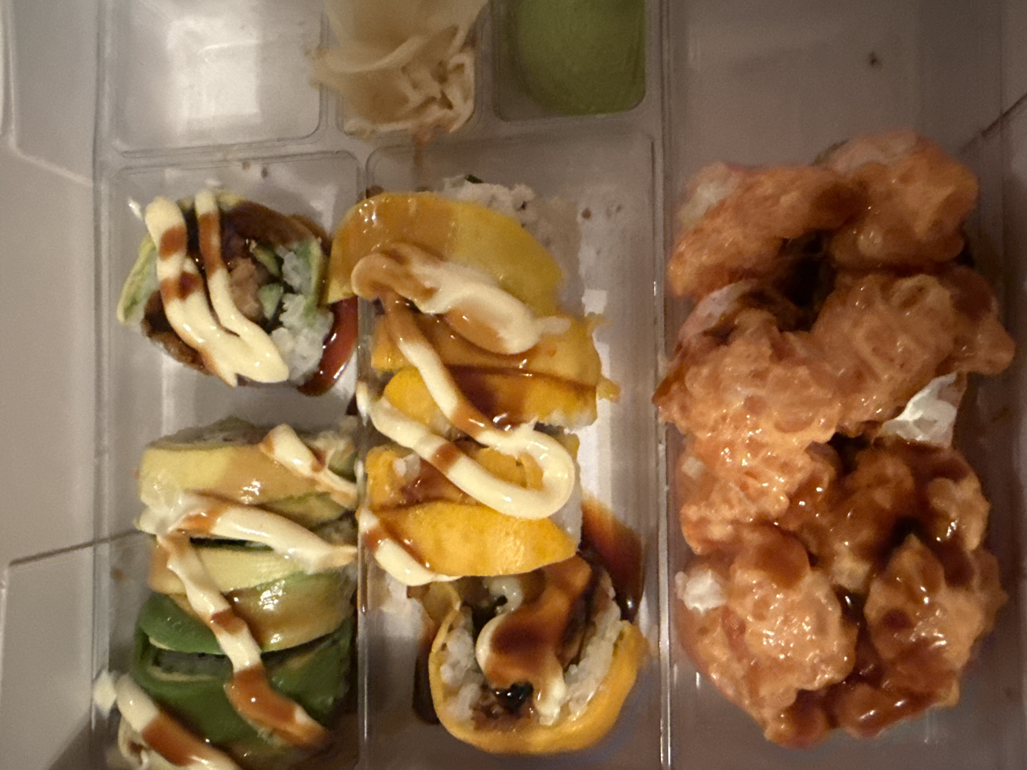 A top-down, close-up shot shows a clear plastic container filled with sushi. The container is divided into sections. In the top left section, a sushi roll with green avocado on the outside, and yellow filling inside is visible, drizzled with white sauce and a darker sauce. To its right, a smaller sushi roll with green avocado, rice, and dark filling, also drizzled with white and dark sauce. In the middle section, a sushi roll with bright yellow mango on the outside, rice, and dark filling is visible, adorned with white sauce and dark sauce drizzles. To the right of the mango sushi, a small compartment contains pickled ginger and a dollop of green wasabi. The bottom section contains a row of sushi rolls covered in a spicy orange sauce, appearing to be fried. The sushi is arranged in a takeaway container. The image does not contain any information indicating its location in Burgas, Bulgaria.