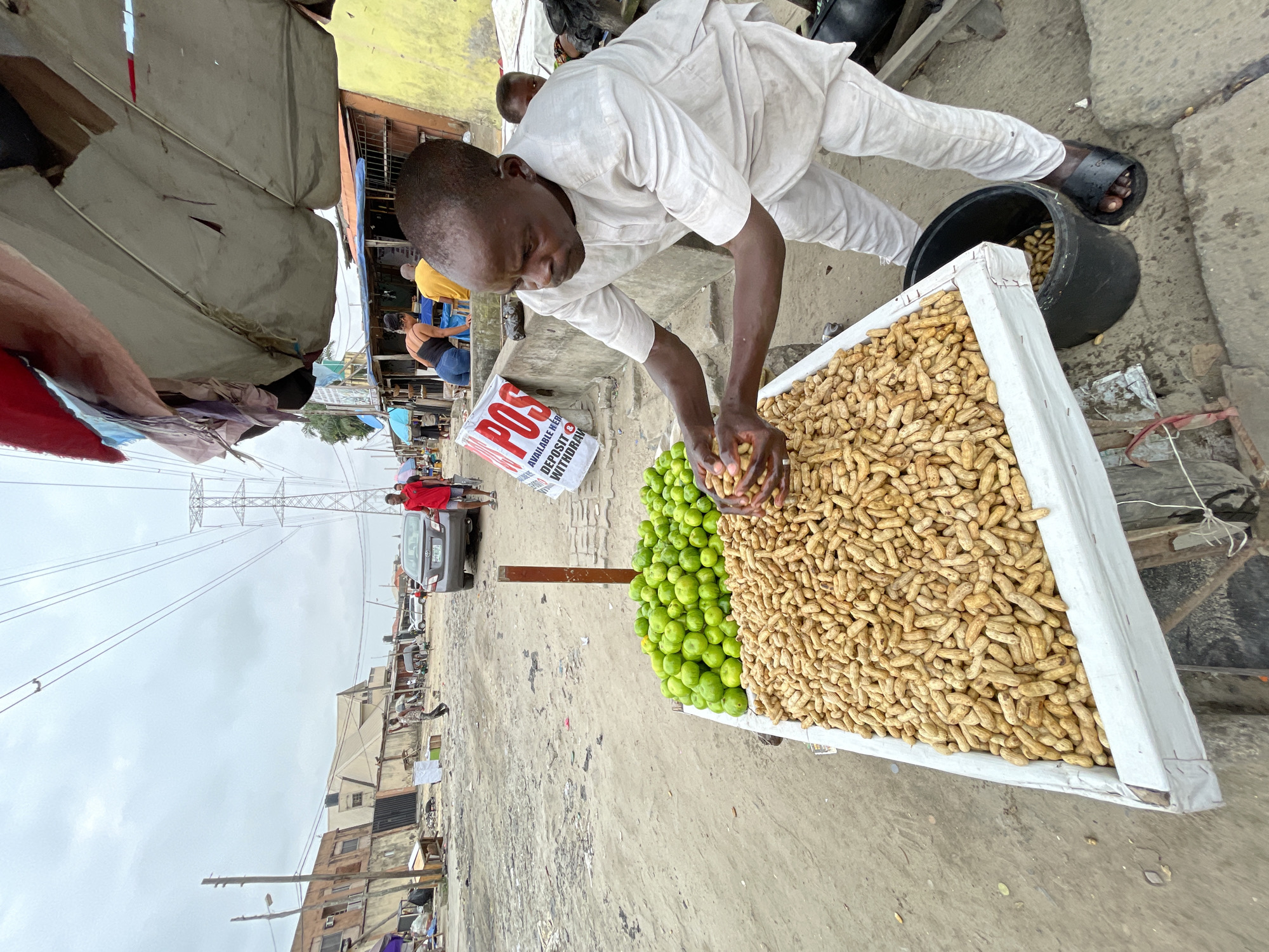 In Ado, Nigeria, a man wearing a light-colored kaftan and dark sandals stands at a market stall, carefully holding a handful of peanuts. He is in the process of transferring the peanuts from the large tray in front of him into a black bucket at his feet. The stall displays a large quantity of peanuts in a white-lined crate, and to the left, a mound of green limes is visible. The ground is sandy and appears to be a dirt road, with debris scattered around. In the background, a grey car is parked, and several buildings line the street. Power lines and a transmission tower are prominent in the sky, indicating an urban or semi-urban setting. A sign reading "POS AVAILABLE HERE DEPOSIT WITHDRAWAL" is visible behind the man. Other people are present in the background, some engaged in their own activities. The scene depicts a typical market day in Ado, Nigeria.