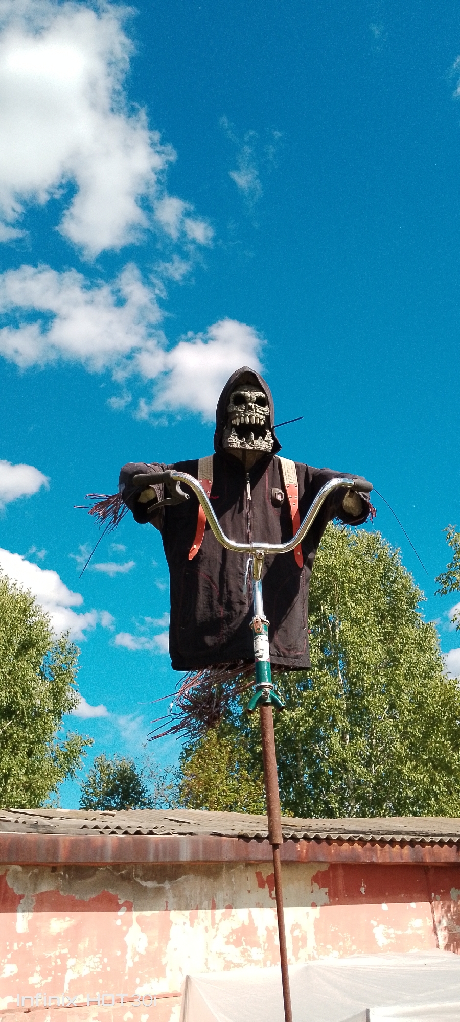 A scarecrow, designed to resemble a grim reaper figure with a skull mask and a dark hooded jacket, is mounted on a pole that extends upwards from behind a low, weathered building. The scarecrow is adorned with red straps and has what appears to be straw or twigs protruding from its arms and below its torso, mimicking the appearance of a tattered garment or broom. The handlebars of a bicycle are integrated into the structure, positioned in front of the scarecrow's torso. The background consists of a bright blue sky with scattered white clouds, framed by the green foliage of trees behind the building and the corrugated metal roof of the structure. The building itself has a predominantly reddish-orange facade with peeling paint and a discolored, aged roof. This scene is located in the city of Kedrovy, Russia, and appears to be in an outdoor, possibly rural or semi-urban, setting.