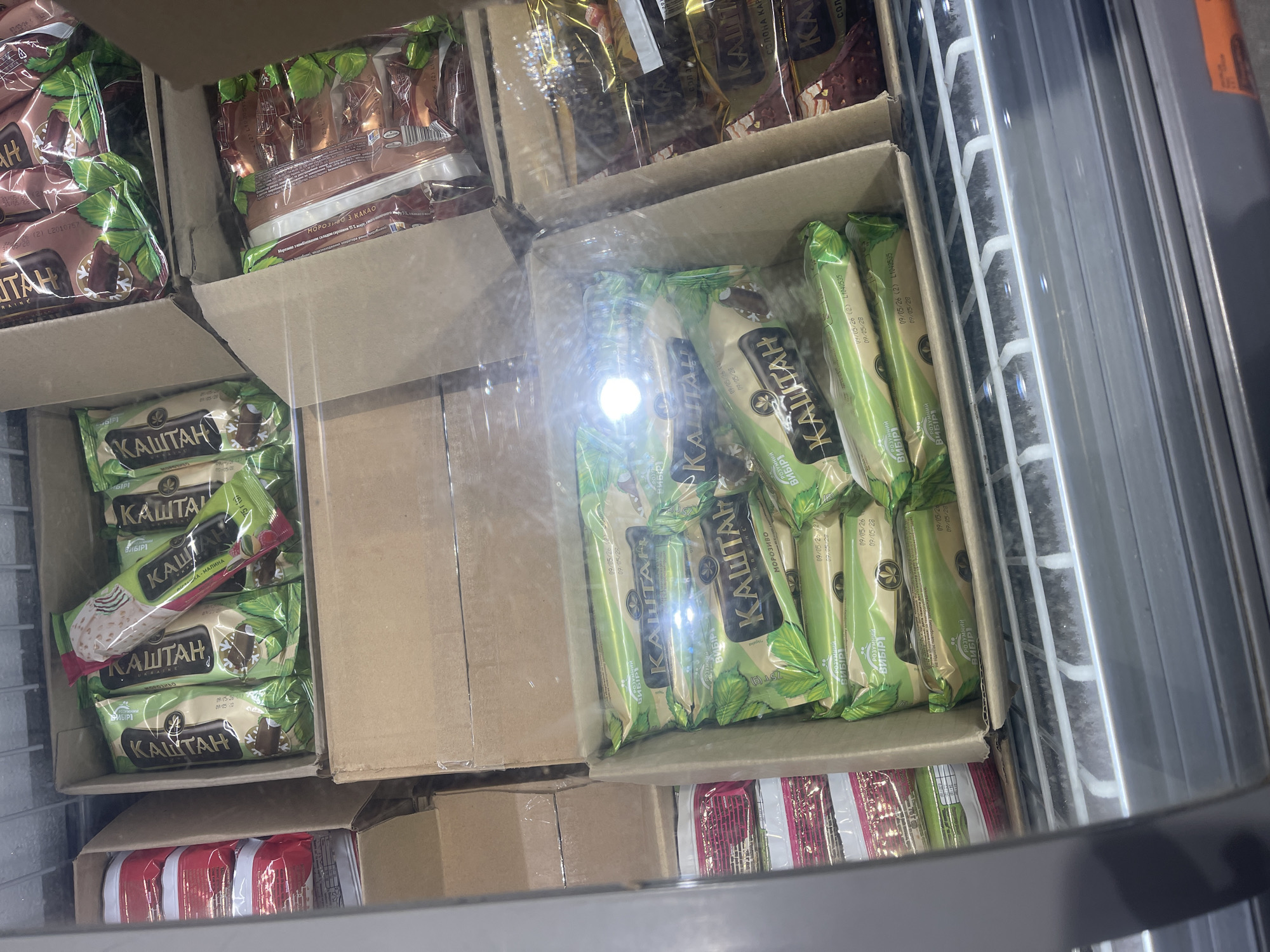 The image shows a top-down view of several boxes of ice cream in a freezer display case. The ice cream bars are individually wrapped in colorful packaging featuring the brand name "КАШТАН" (Kashtan). Some packages are green and white, while others are brown and yellow. The ice cream bars appear to be of different flavors, with some showing images of chocolate or nuts. The boxes are stacked inside the freezer, and some are partially obscured by others. The freezer display case has metal shelving, and the overall environment is cold, as indicated by the frost on the shelves. There are no people visible in the image. The location is stated to be Unknown, Unknown.