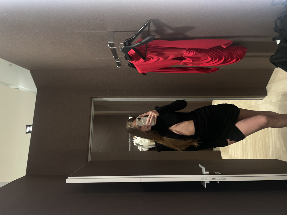 In a dressing room, a person wearing a black dress takes a mirror selfie with a smartphone. The reflection shows the person from the chest down. A red dress hangs on a rack above. The dressing room features brown walls and a door with a prominent silver handle. The floor is light-colored and patterned. The person's phone case is white.
