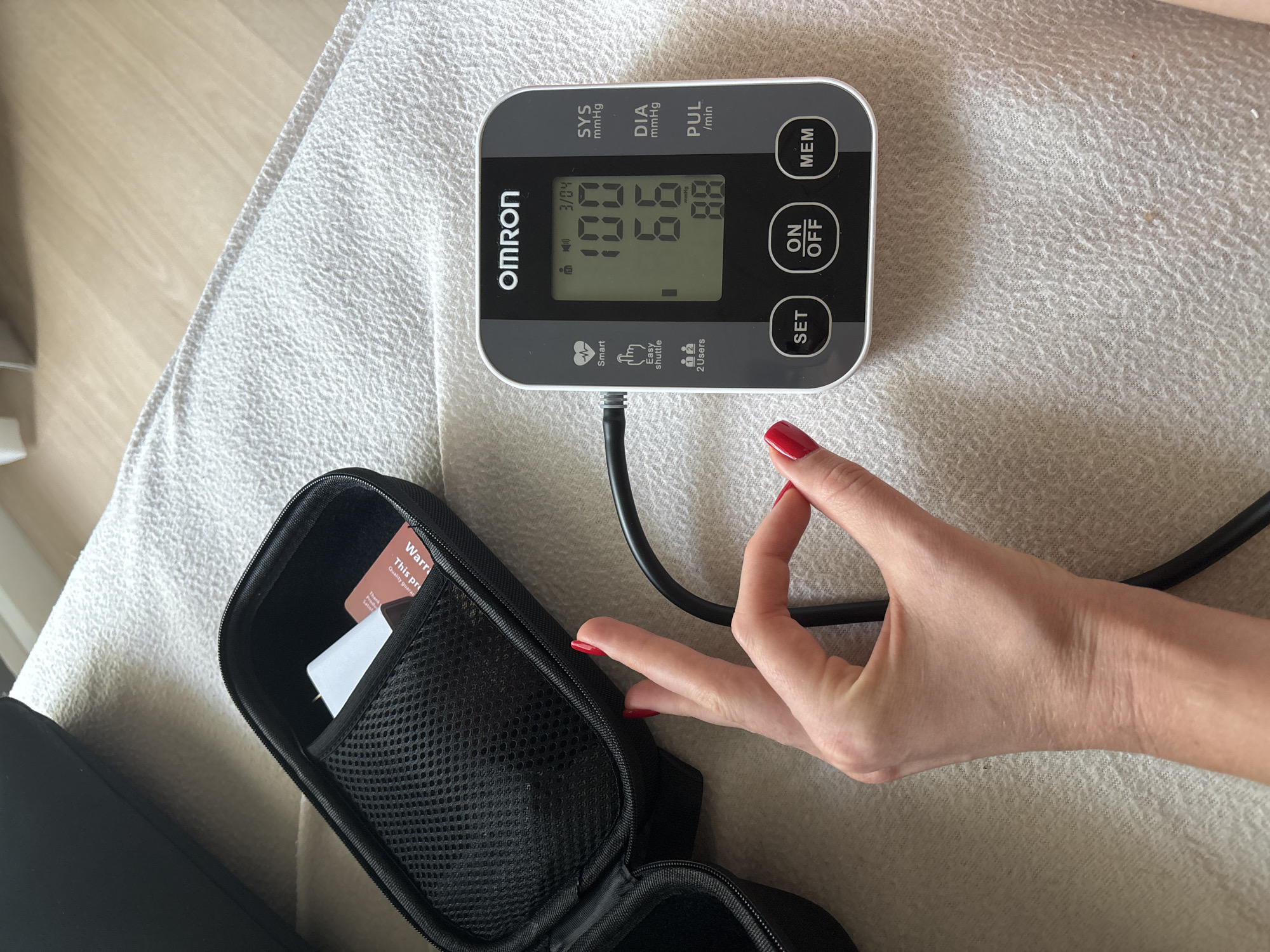 A blood pressure monitor, a black zippered case with cards inside, and a woman's hand are on a textured surface that appears to be a couch. The blood pressure monitor is a gray and white Omron device with the readings "100," "59," and "88" displayed on its screen. The date 3/04 is also visible. The woman's hand with red nail polish is near the monitor's black tube. The black case contains a card with text "Warra" and "Quality guaran" partially visible. The environment suggests an indoor setting on a couch with neutral color fabric. There is no indication of this image being taken in Choeng Thale, Thailand.
