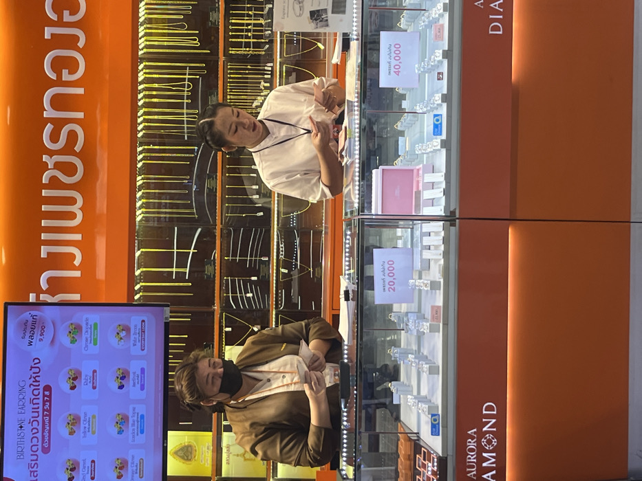 This image depicts the interior of a jewelry store, identified as an Aurora Diamond and Gold Shop ("ห้างเพชรทองออโรร่า"), located in Bang Pu Subdistrict Municipality, Thailand. Two adult female employees are positioned behind a glass display counter. The employee on the left wears a brown jacket over a light shirt and a black face mask, with her dark hair pulled back. She is counting or handling small slips of paper or currency. The employee on the right wears a white short-sleeved collared shirt, with her dark hair styled in braids, and she is also counting or handling similar items.
The counter features two illuminated glass display cases containing various rings on white stands, with some price tags visible (e.g., "21,900"). White signs within the cases indicate "20,000" and "40,000" in Thai script. A pink jewelry box is present in the right display. The base of the counter is orange, displaying "AURORA DIAMOND" branding.
In the background, behind the employees, a large wall display unit is filled with numerous gold necklaces and chains hanging vertically. Above this, a prominent orange panel features the store's name in white Thai script. To the upper left, a digital screen displays a "BIRTHSTONE EARRING" promotion, showing various gemstone options and a price of "9,900." The environment is brightly lit and organized, characteristic of a retail jewelry establishment.