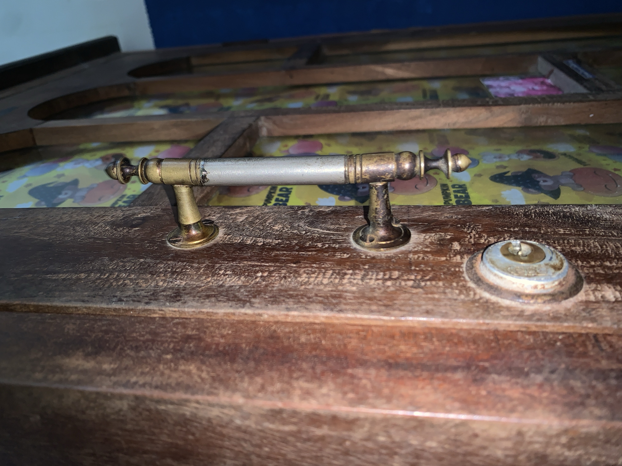 The image shows a close-up, eye-level, side-view of a wooden door with a brass handle and lock. The door has a weathered, textured surface with vertical wood grain. The brass handle is ornate, with decorative ends and a cylindrical central grip that appears to be silver-toned with some wear. Below the handle is a circular brass lock mechanism, showing signs of age and use. Behind the door, a colorful pattern with cartoon bears wearing witch hats is visible, suggesting the door might lead to a themed room or storage area. The background shows the edge of a wooden cabinet or frame and a dark blue wall. The scene is lit from the side, casting shadows and highlighting the textures of the wood and metal. The image does not provide any information about the city of Karangkemiri, Indonesia, nor does it depict any people, actions, or interactions.